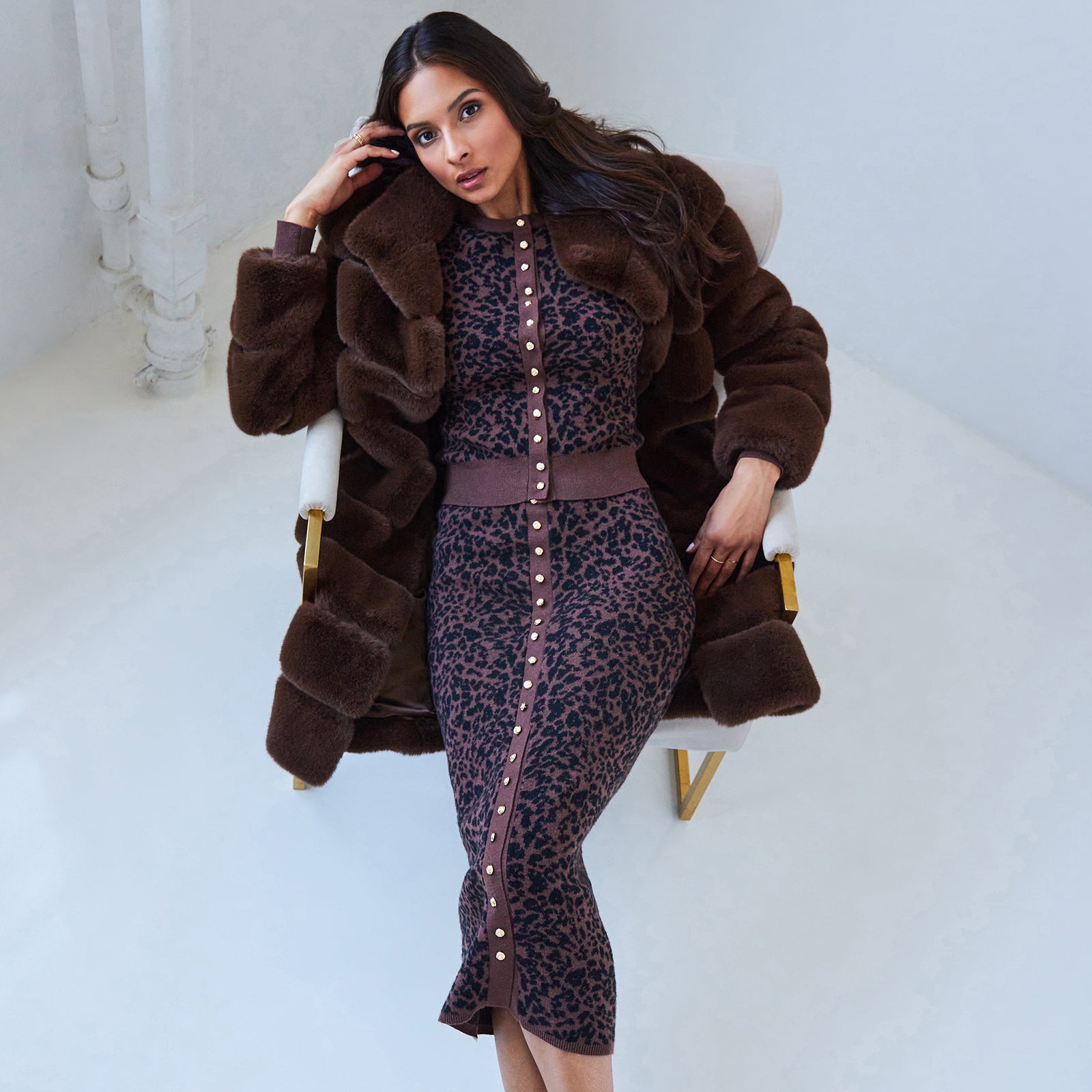 Woman wearing a brown fur coat and leopard print outfit on a white chair.