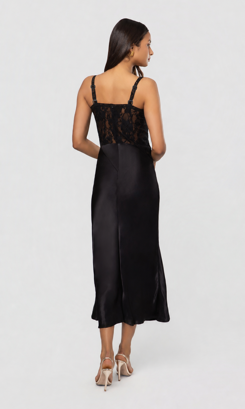 Back view of woman wearing a black satin slip dress with lace detailing on a white background
