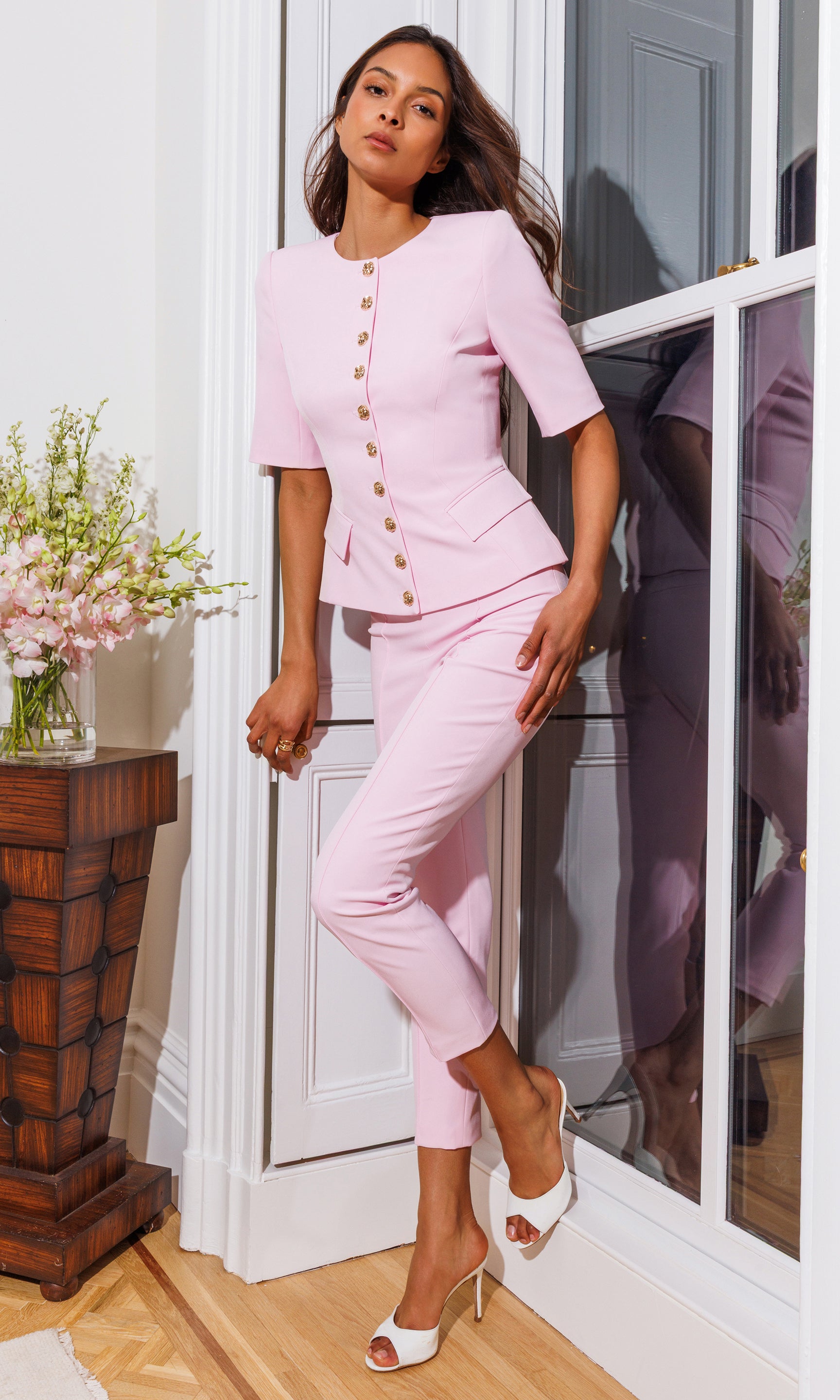 Model wears the Lonny Crepe Pants in soft rose in an editorial full body view by the window, styled with heels and a matching pink jacket for a polished, feminine look.