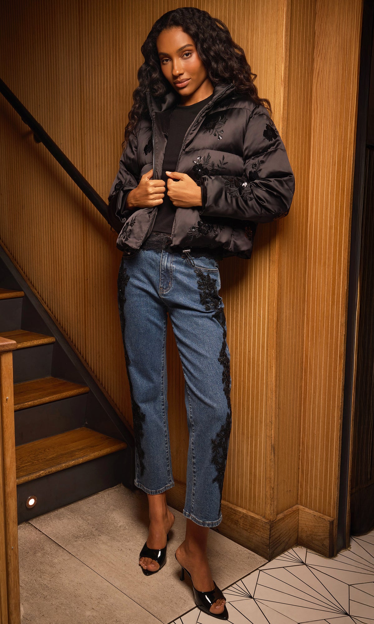 Model wearing a black quilted puffer jacket, black top, blue jeans with black lace applique, and black heeled sandals.
