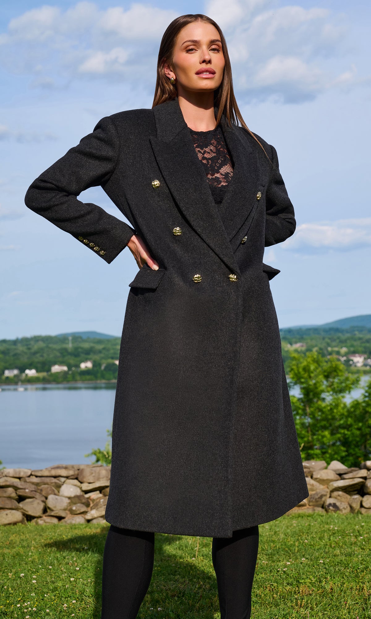 A woman stands outdoors wearing a long double-breasted charcoal gray coat with gold-tone buttons over a black lace top and black leggings.