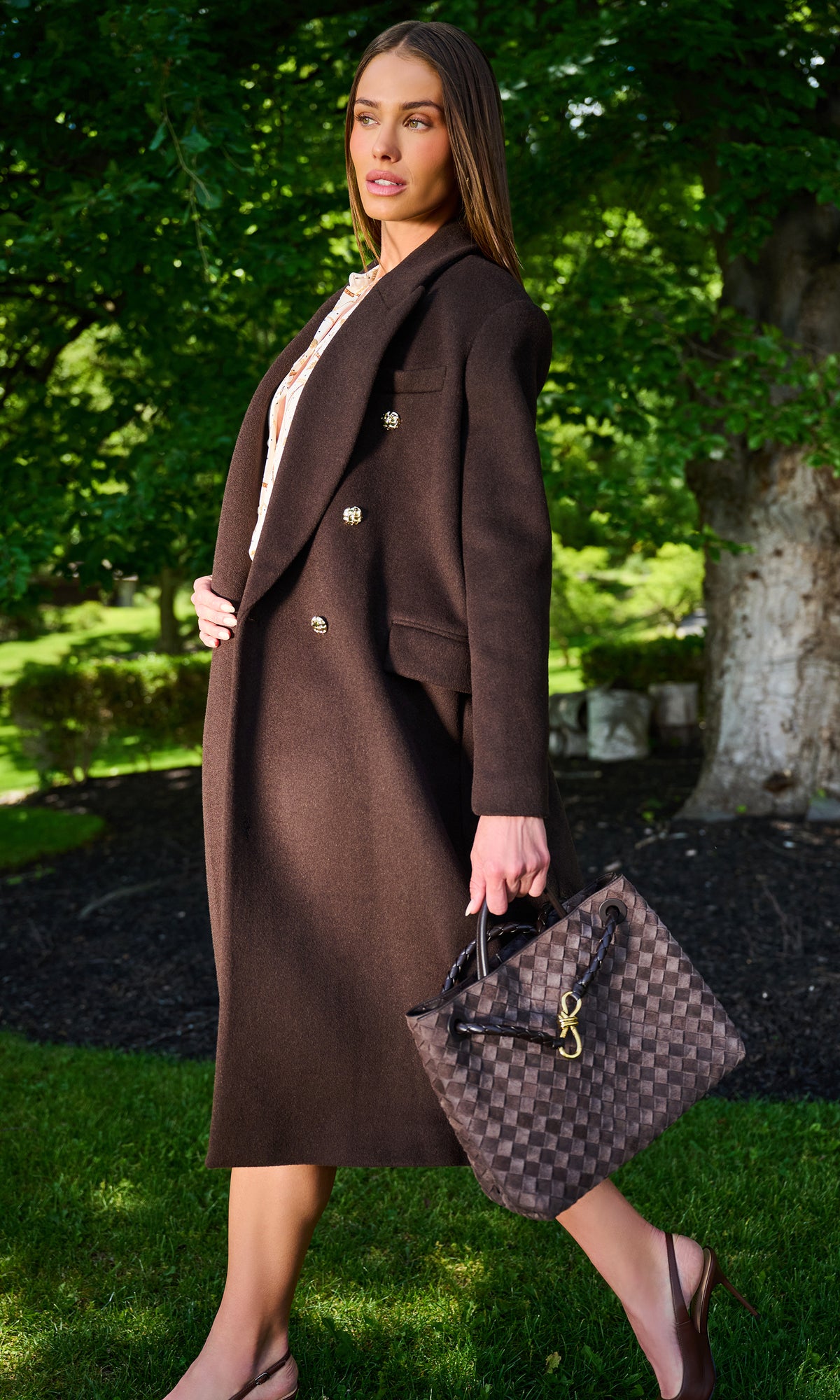 A woman stands outdoors on a grassy lawn in front of a tree, wearing a long brown double-breasted coat and holding a brown checkered handbag.