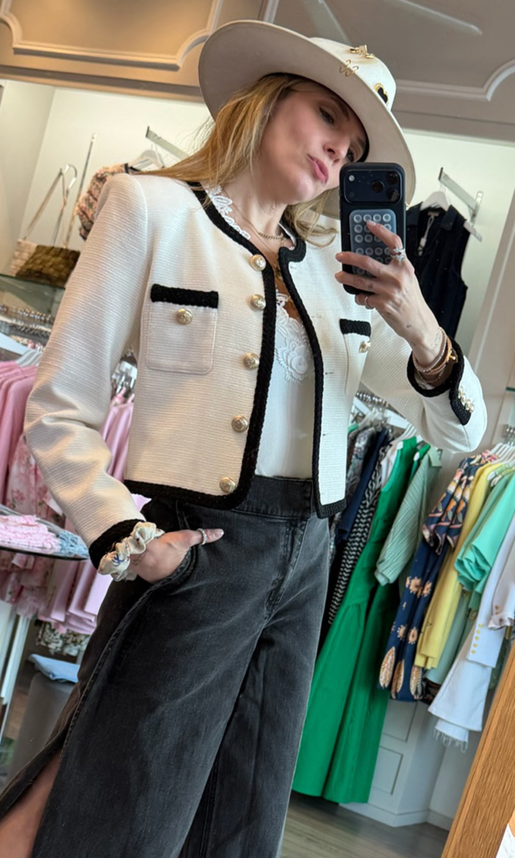Founder Roni taking a mirror selfie wearing a white jacket with black trim, black pants, and a white hat in a clothing store.