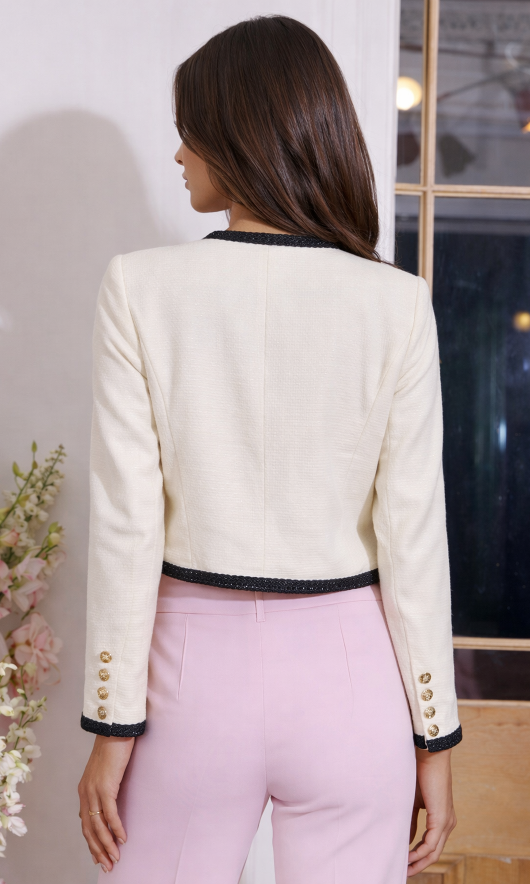 Back view of woman wearing a cream blazer with black trim and pink pants in an indoor setting.