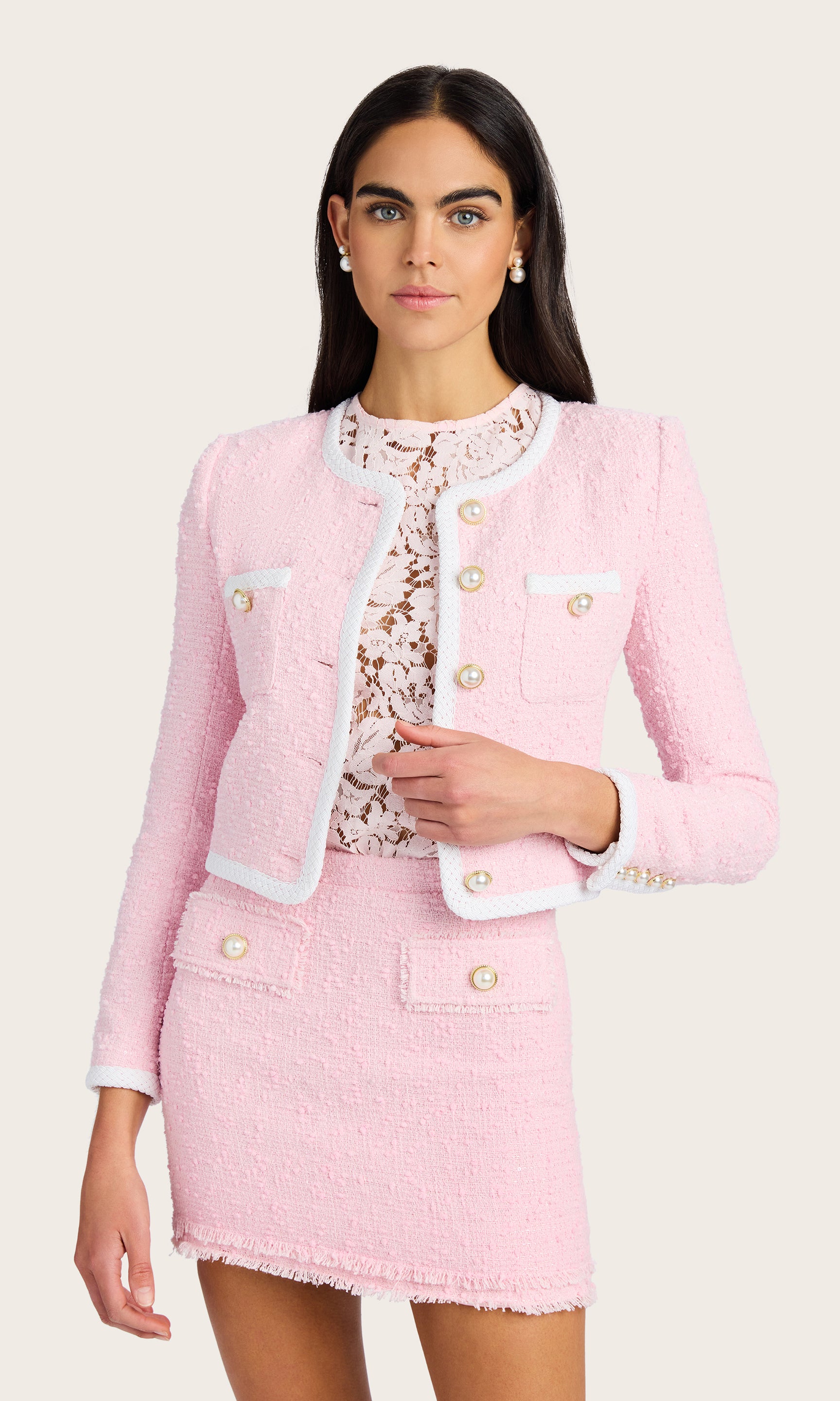 front view of model wearing a light pink Amber tweed jacket with pearl buttons, paired with a matching light pink tweed skirt, and a light pink lace under shirt, accessorized with pearl earrings