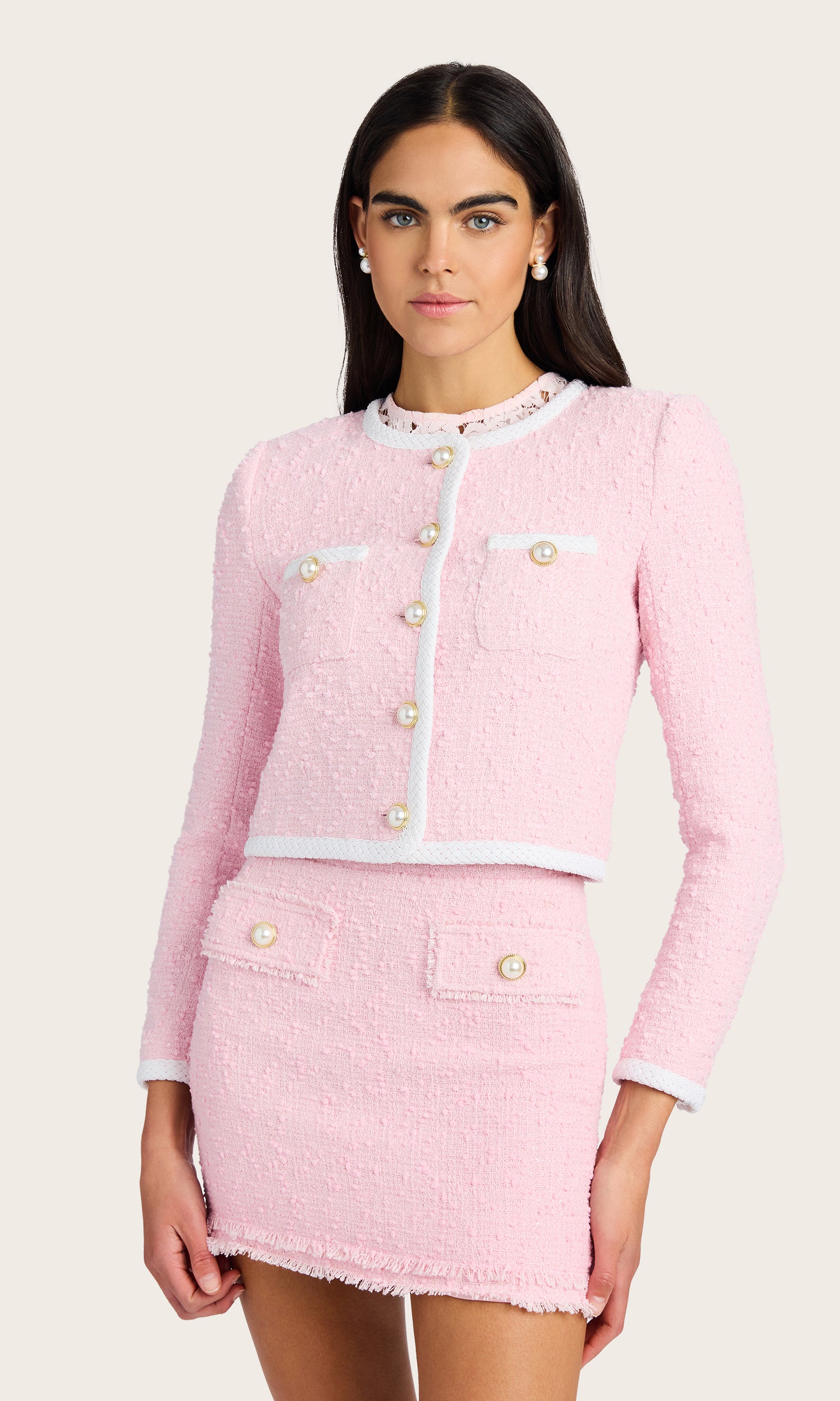 3/4 view of model wearing a light pink Amber tweed jacket with pearl buttons, paired with a matching light pink tweed skirt, and a light pink lace under shirt, accessorized with pearl earrings