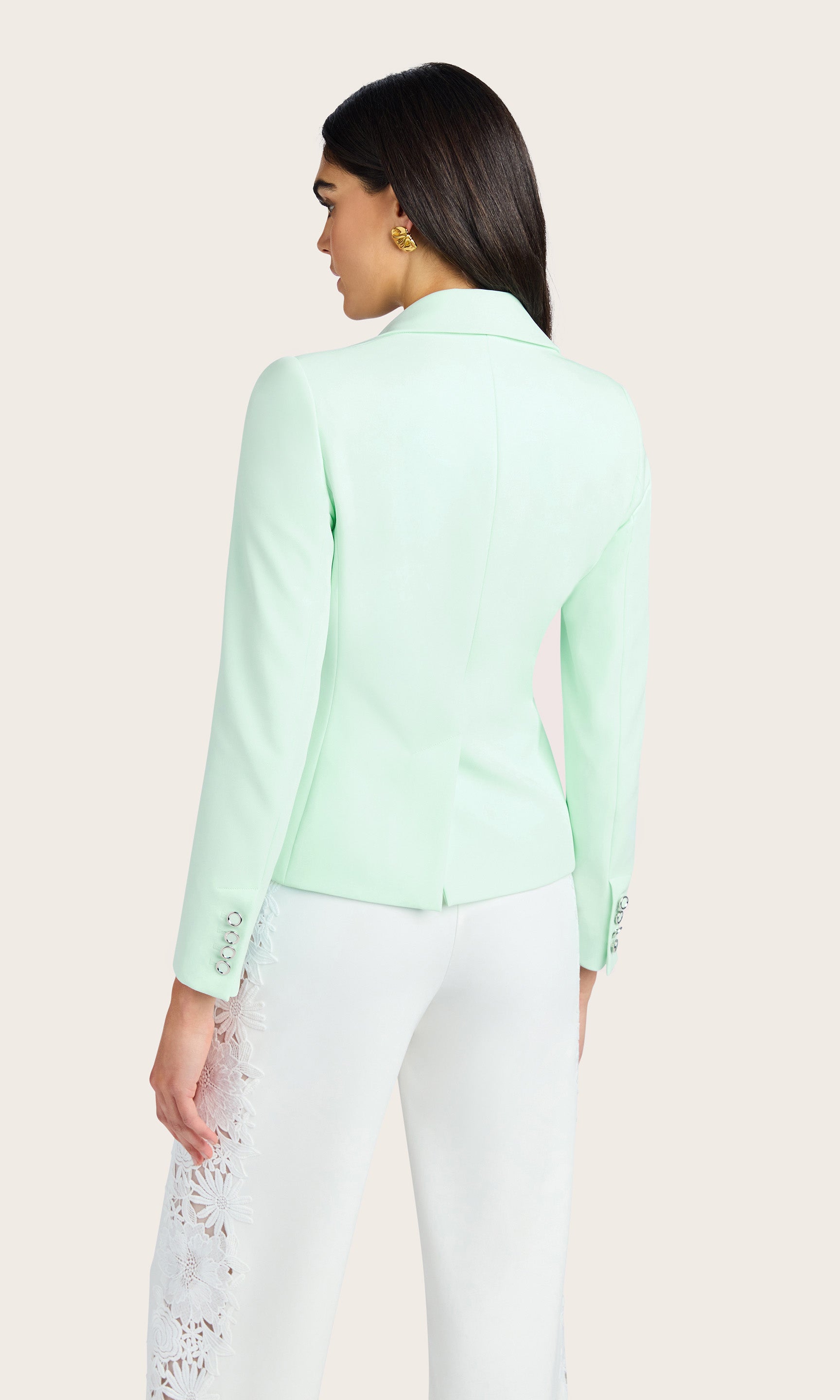 back view of model wearing a crepe seafoam double breasted blazer, paired with white crepe pants with side lace details, accessorized with a gold earrings