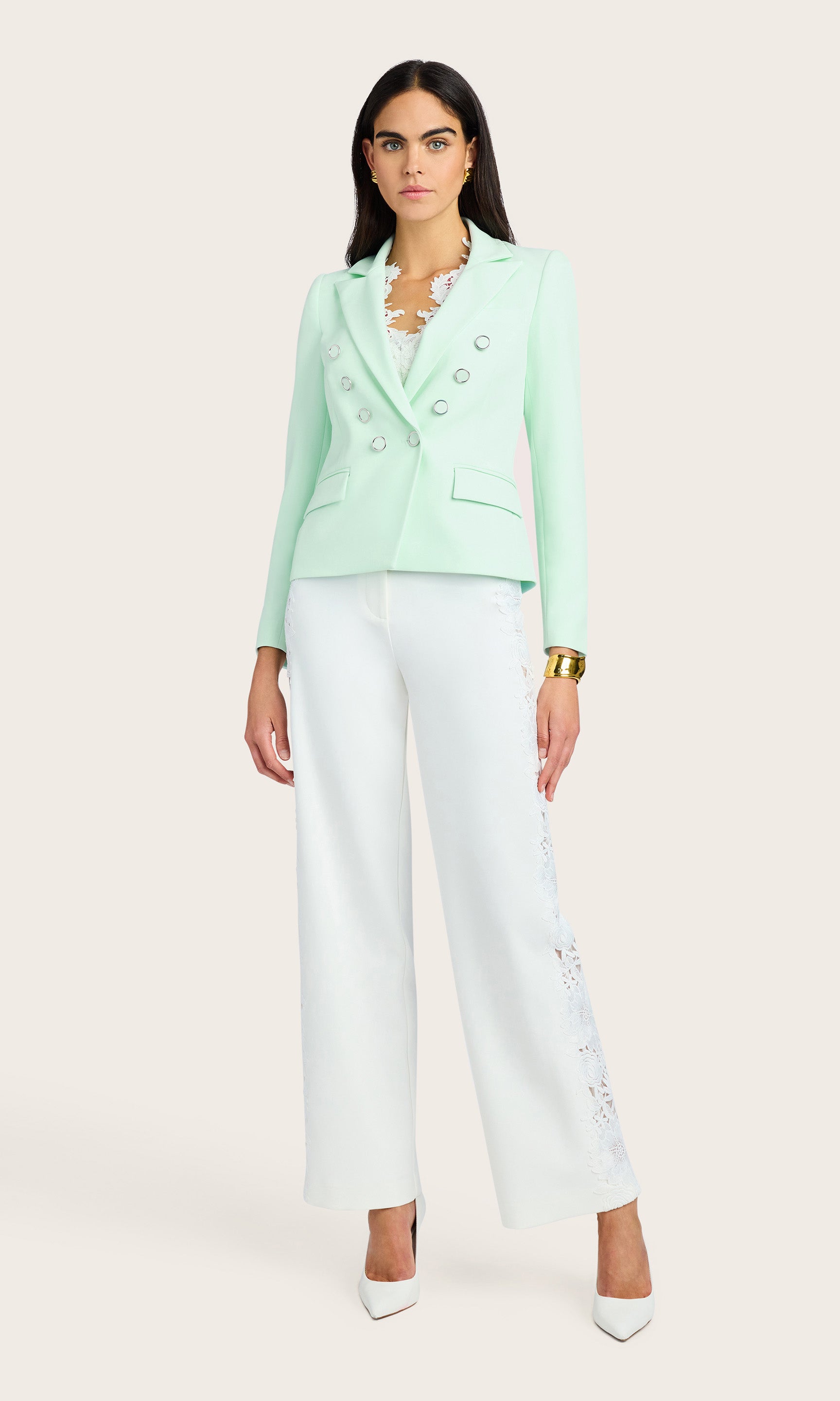 full body view of model wearing a crepe seafoam double breasted blazer with silver adorned buttons, paired with white crepe pants with side lace details, accessorized with a gold bracelet, earrings and white kitten heels