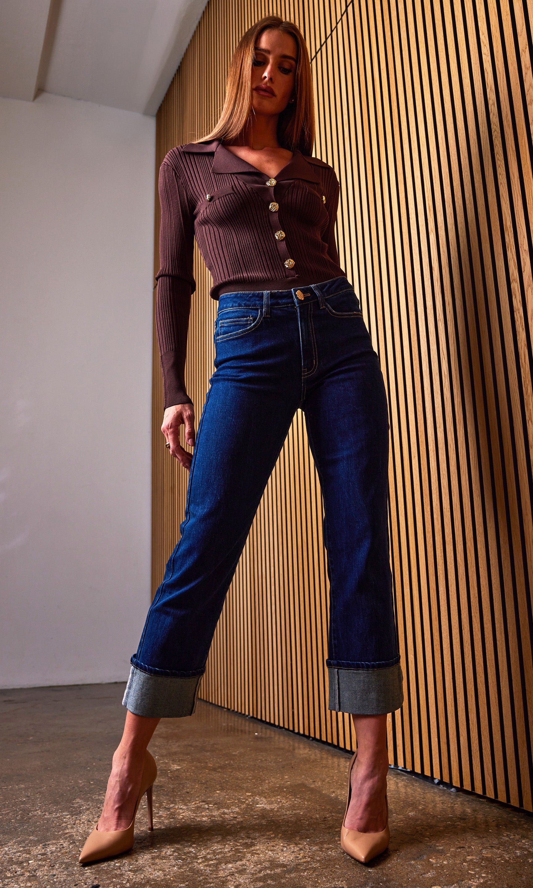 Woman wearing a brown ribbed top and blue jeans standing against a wooden paneled wall.
