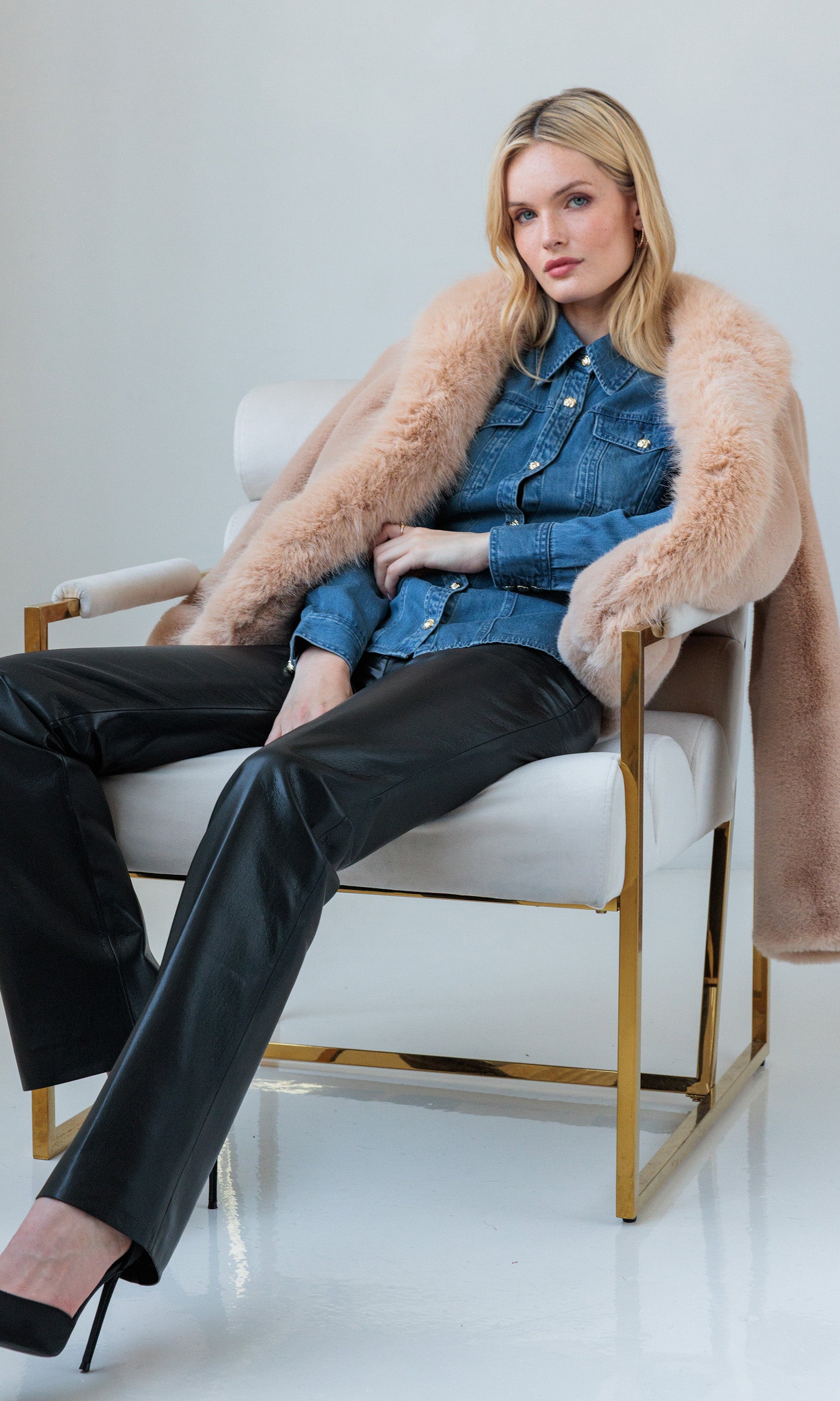 Seated full body view of the model in the Antoinette Denim Shirt paired with black pants and a faux fur coat draped over the chair.