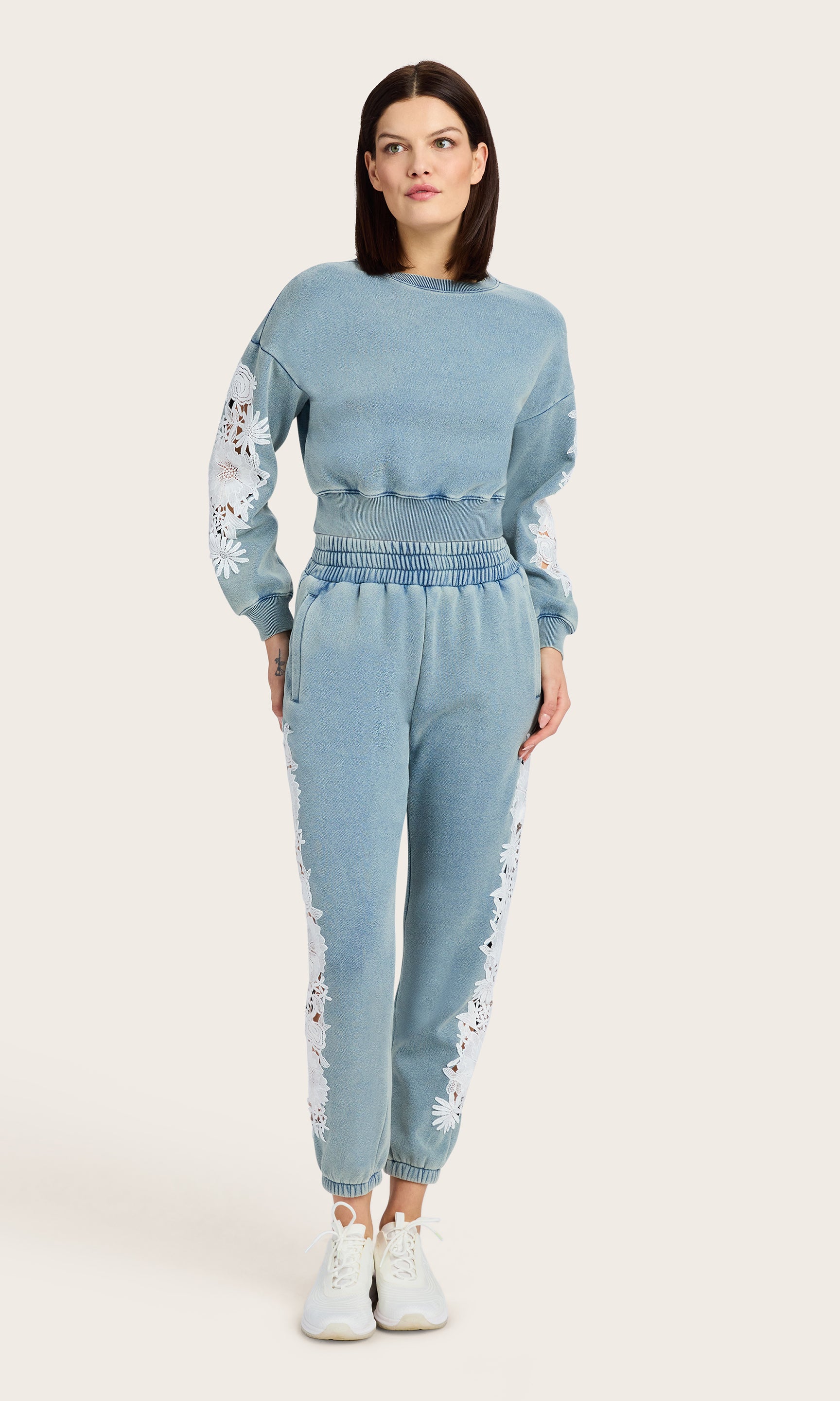 full body image of model wearing the Arabella washed denim sweatshirt with white lace details on the sleeves, paired with matching washed denim joggers and white tennis shoes