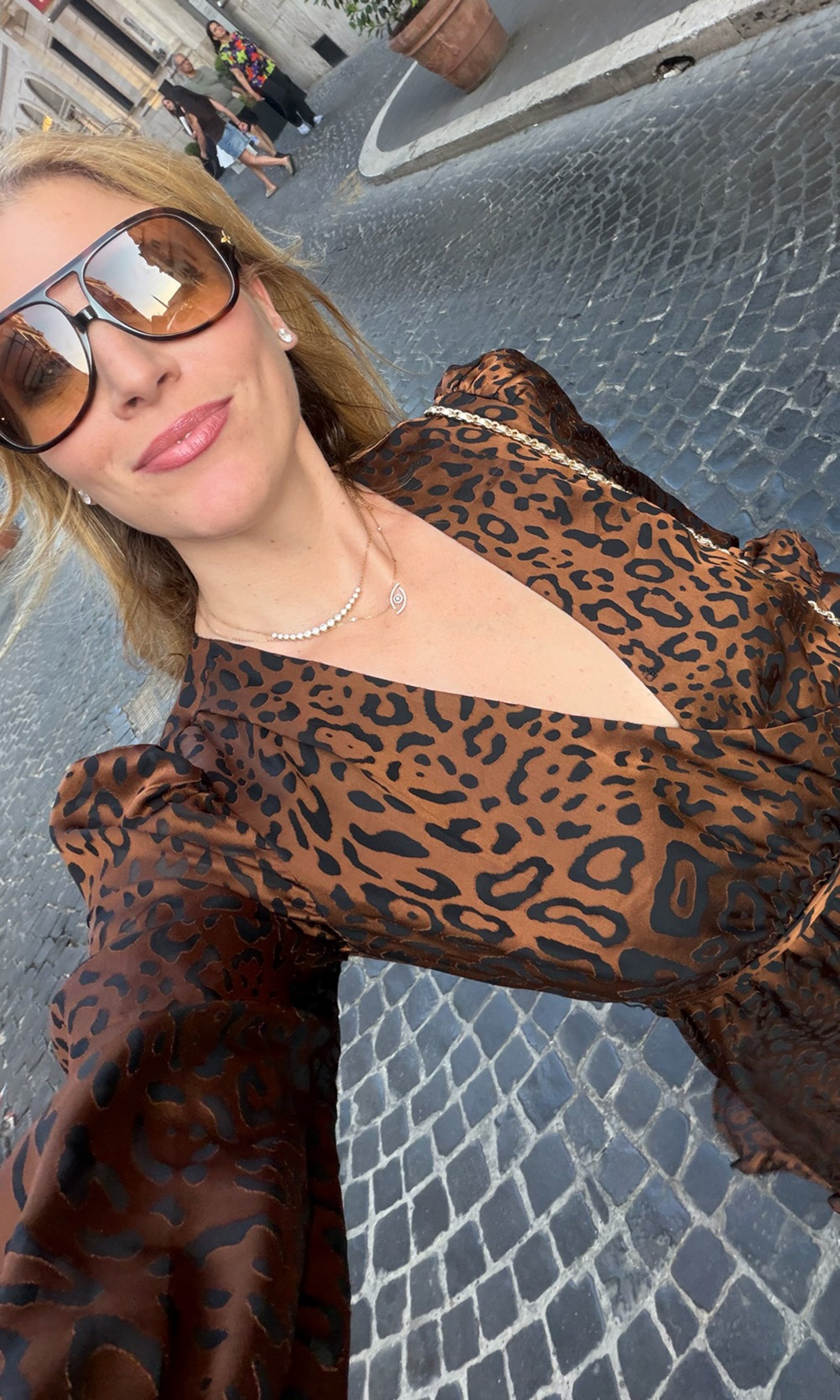 Founder taking a selfie outdoors on a cobblestone street, wearing a brown leopard-print top with a deep V-neckline and oversized sunglasses.
