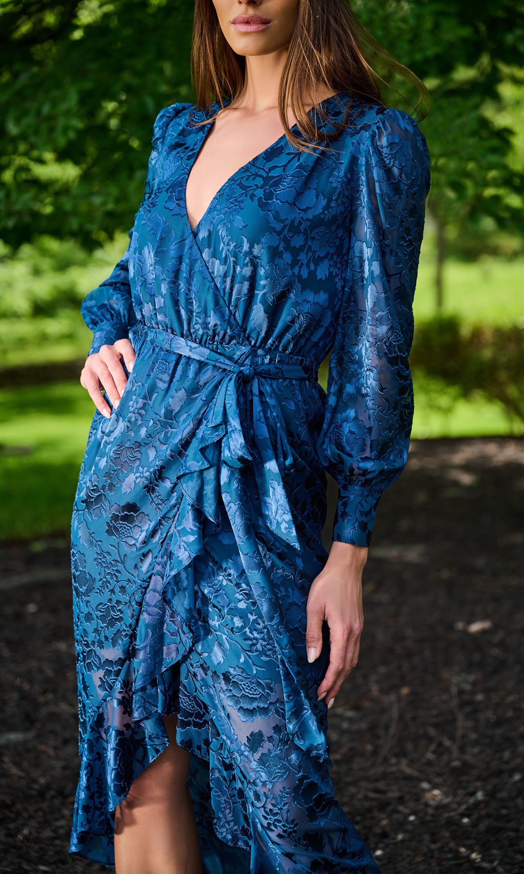 Model wearing a blue patterned midi dress with long puff sleeves, and a matching waist tie, photographed outdoors in a park setting.