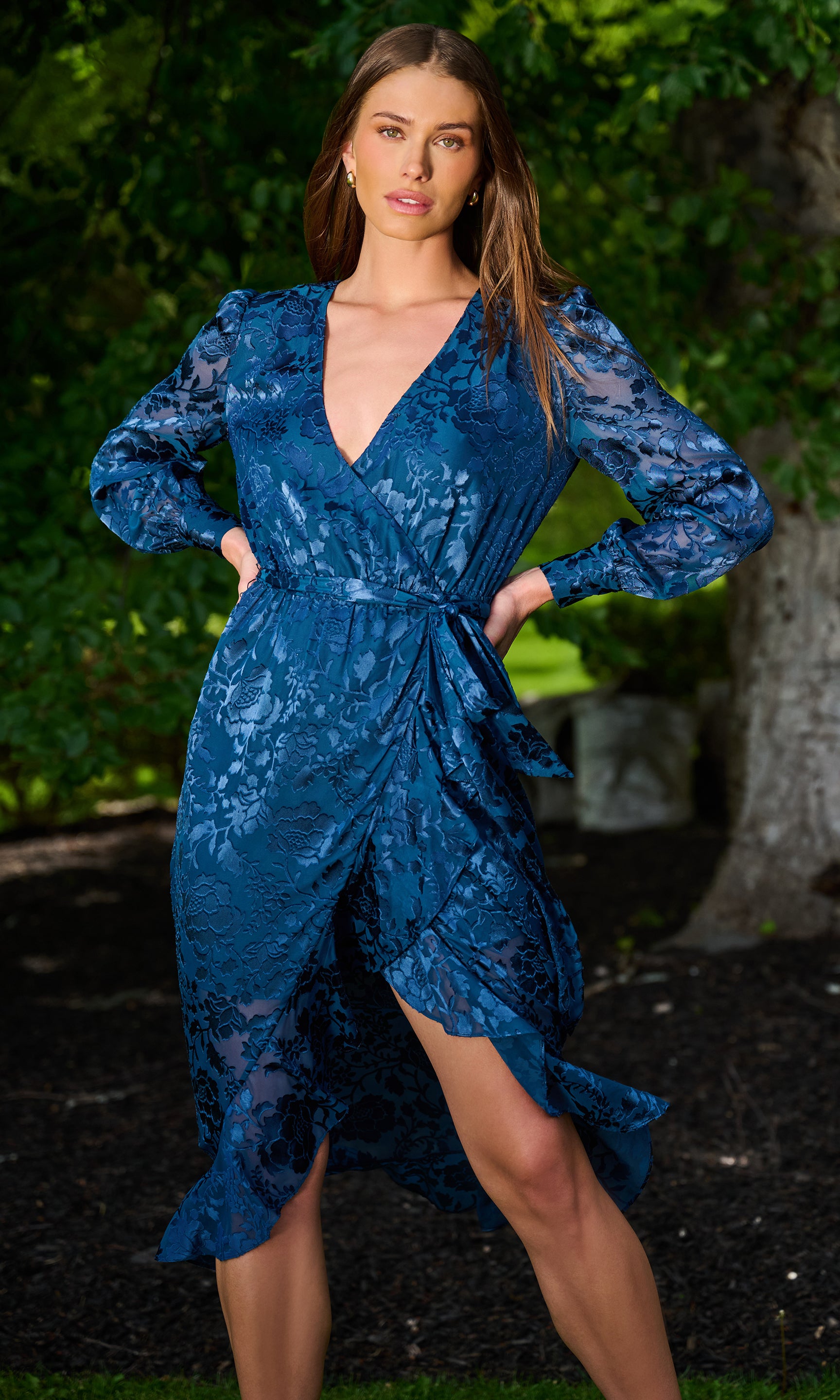 A woman wearing a blue lace wrap dress with long sleeves and a V-neckline, standing outdoors in front of a tree with green foliage.