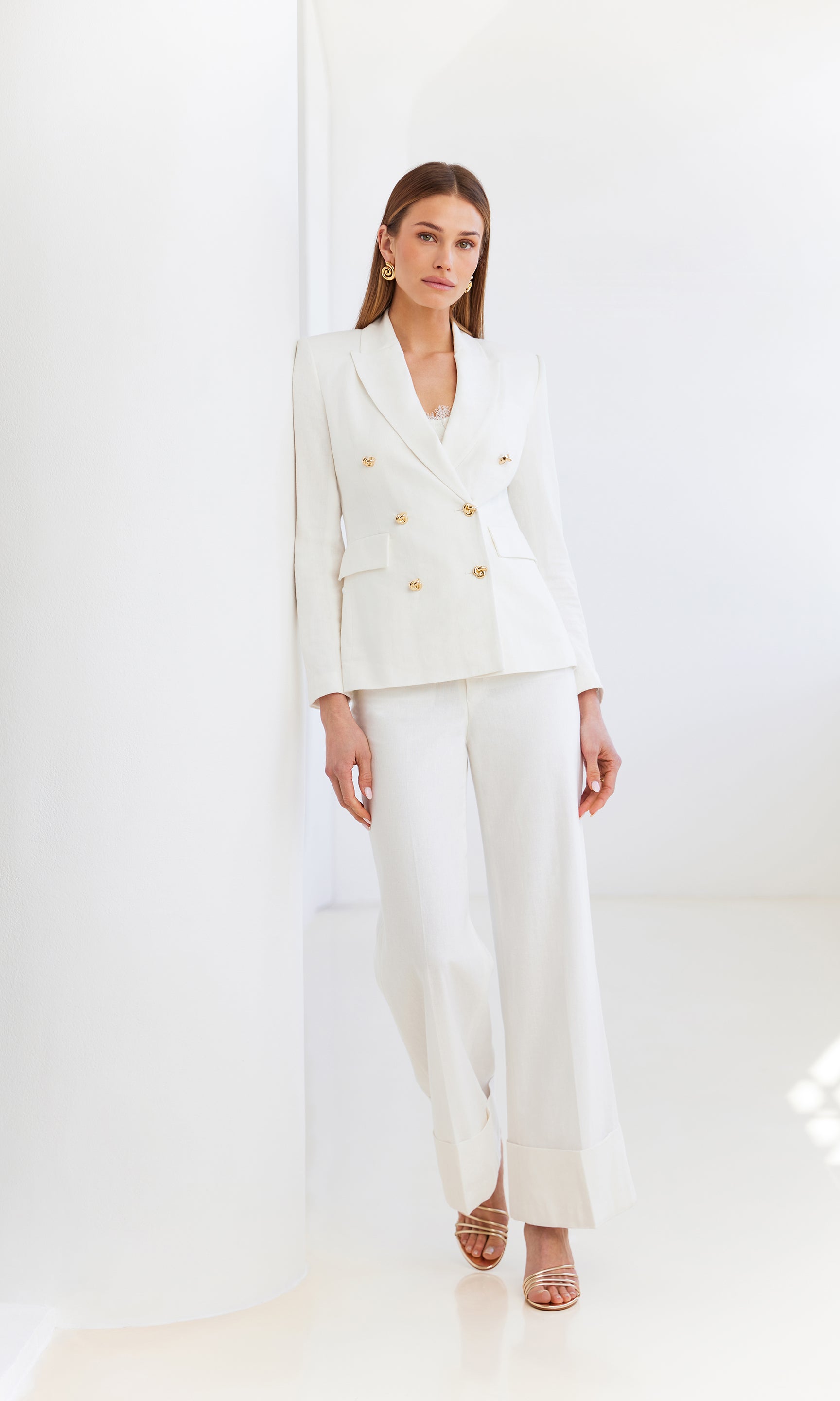 image of model leaning against a white wall wearing the white Arielle blazer with matching white trousers and silver heels