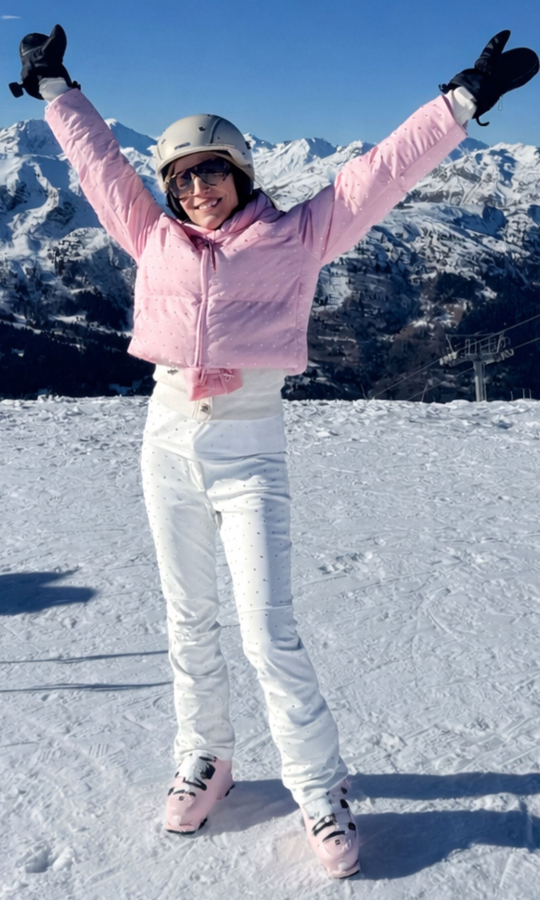 Person in pink jacket and white pants standing on a snowy slope with mountains in the background