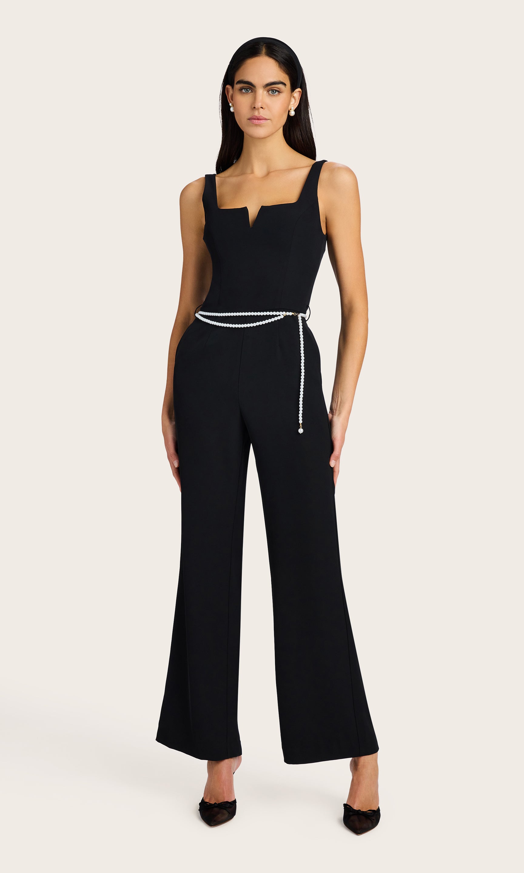 front view of model wearing the Balius light weight crepe jump suit in black with a white pearl belt, accessorized with black kitten heels and pearl earrings