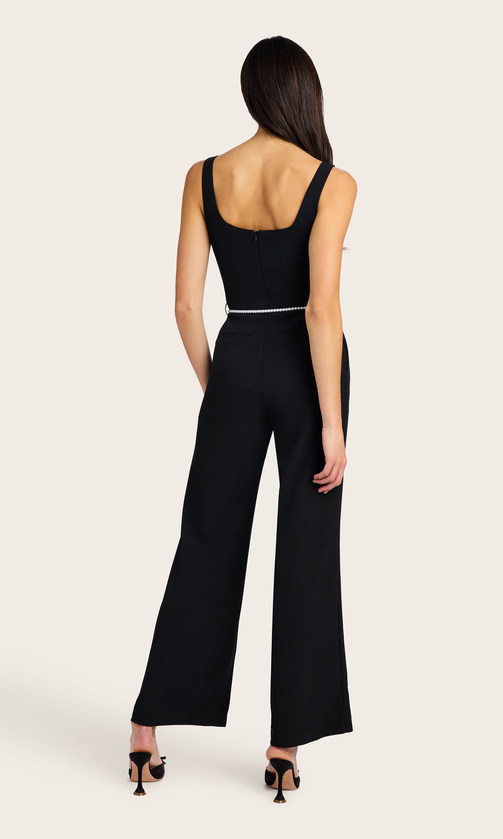 back view of model wearing the Balius light weight crepe jump suit in black with a white pearl belt, accessorized with black kitten heels and pearl earrings