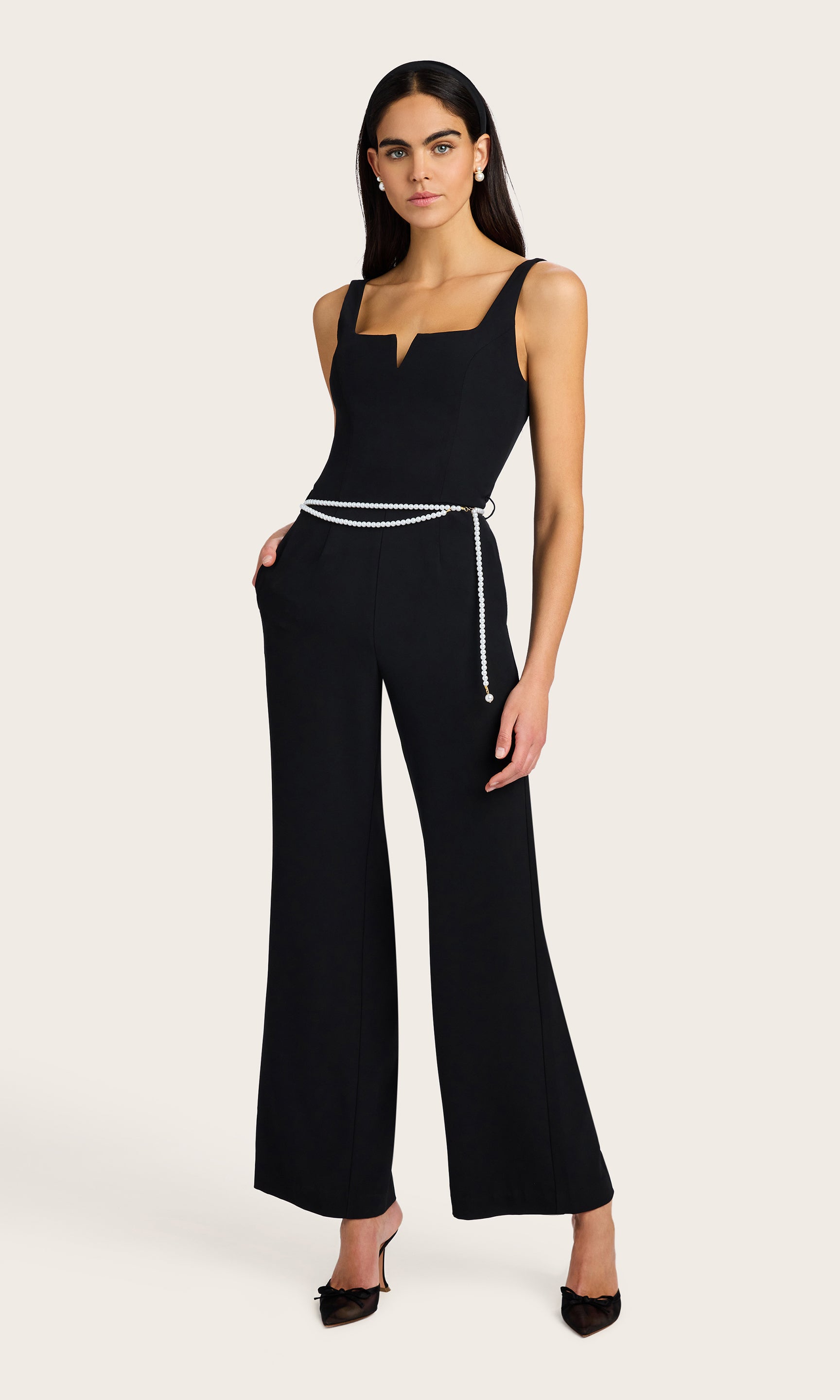 3/4 front view of model wearing the Balius light weight crepe jump suit in black with a white pearl belt, accessorized with black kitten heels and pearl earrings