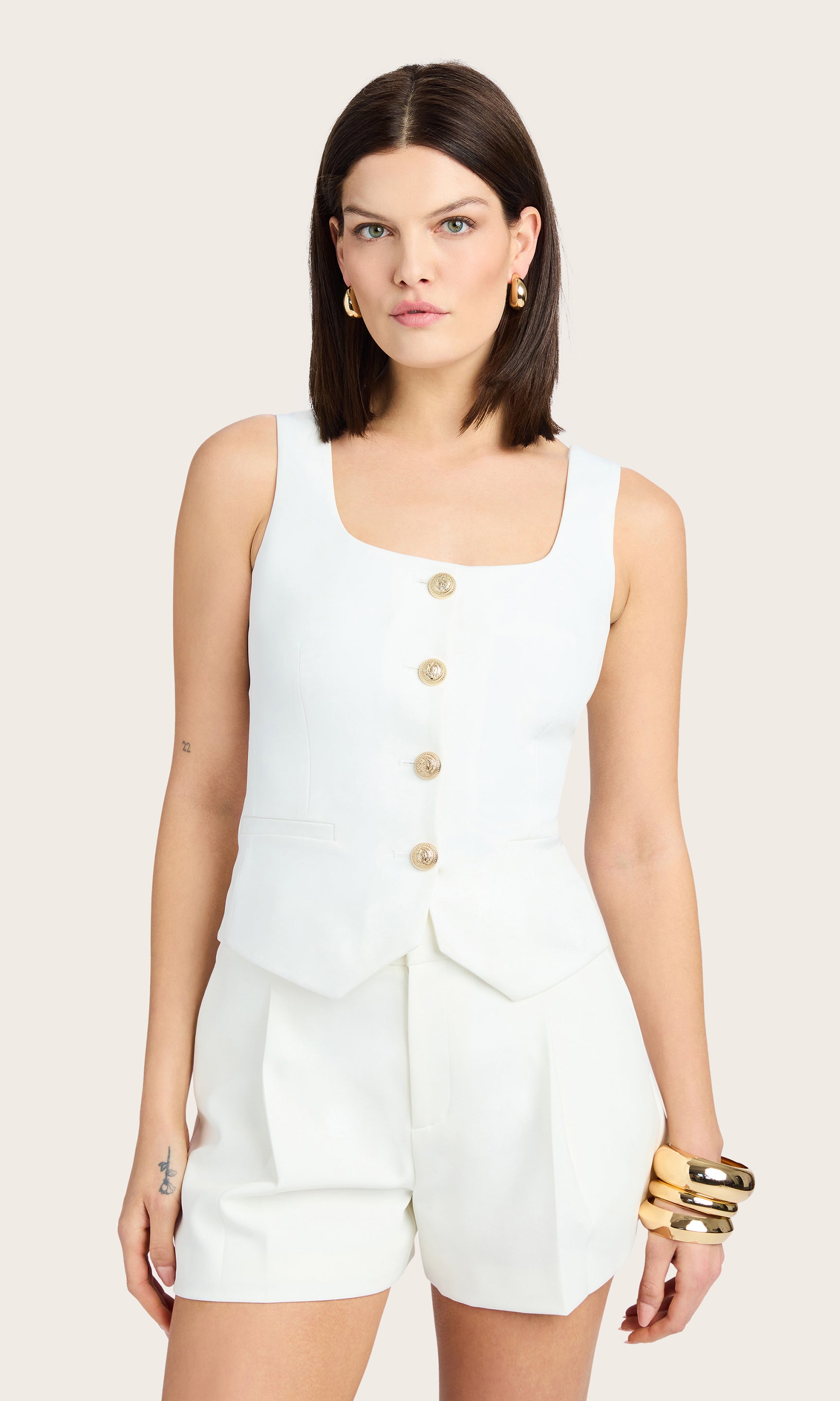 front view of model wearing Beatriz top in white with ripple gold buttons down the front, paired with matching white crepe shorts and accessorized with a gold bracelet and earrings