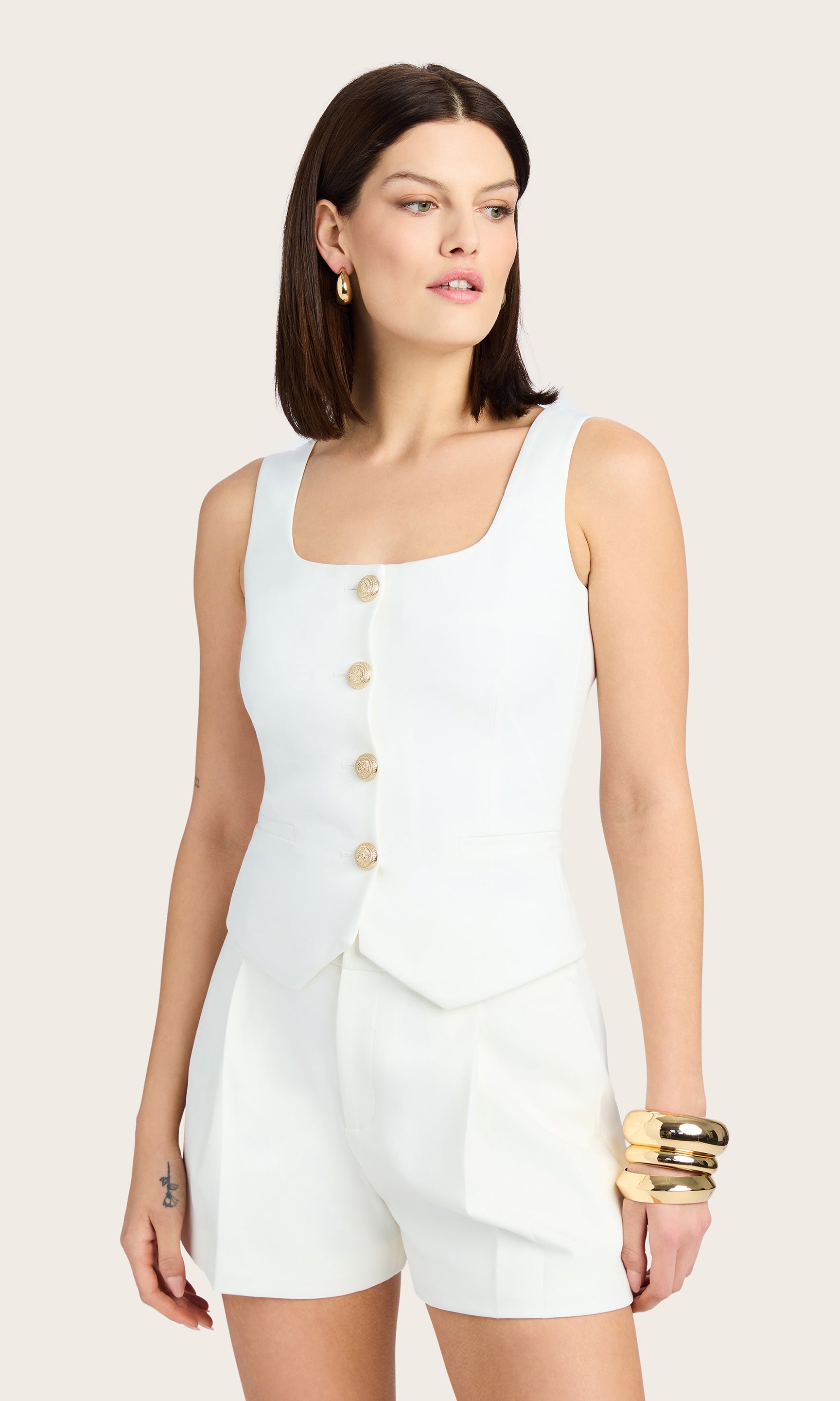 3/4 front view of model wearing Beatriz top in white with ripple gold buttons down the front, paired with matching white crepe shorts and accessorized with a gold bracelet, earrings