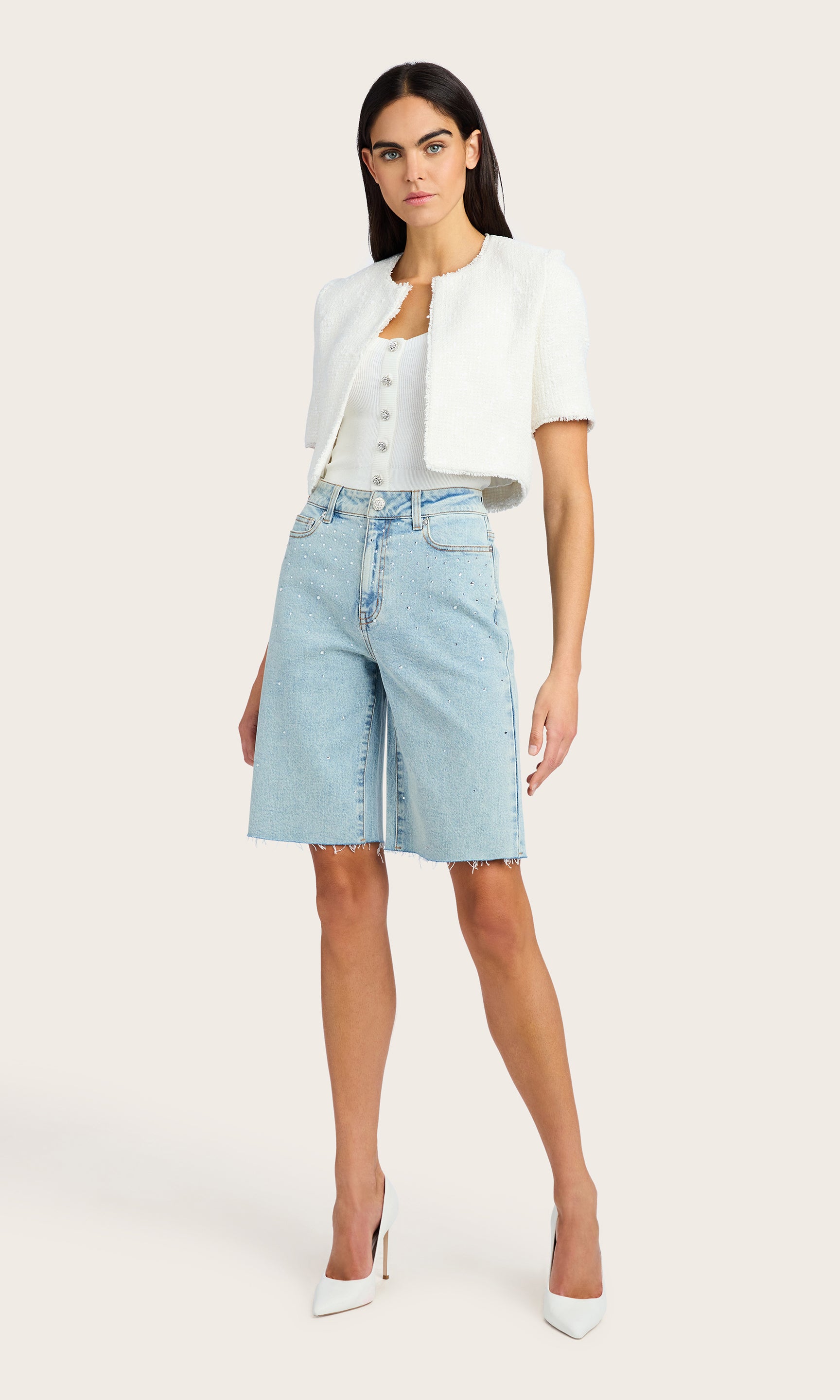 full body image of model wearing the Bellina bermuda shorts in light blue denim adorned with clear crystals, paired with a white short sleeve tweed jacket over a white tank and white kitten heels