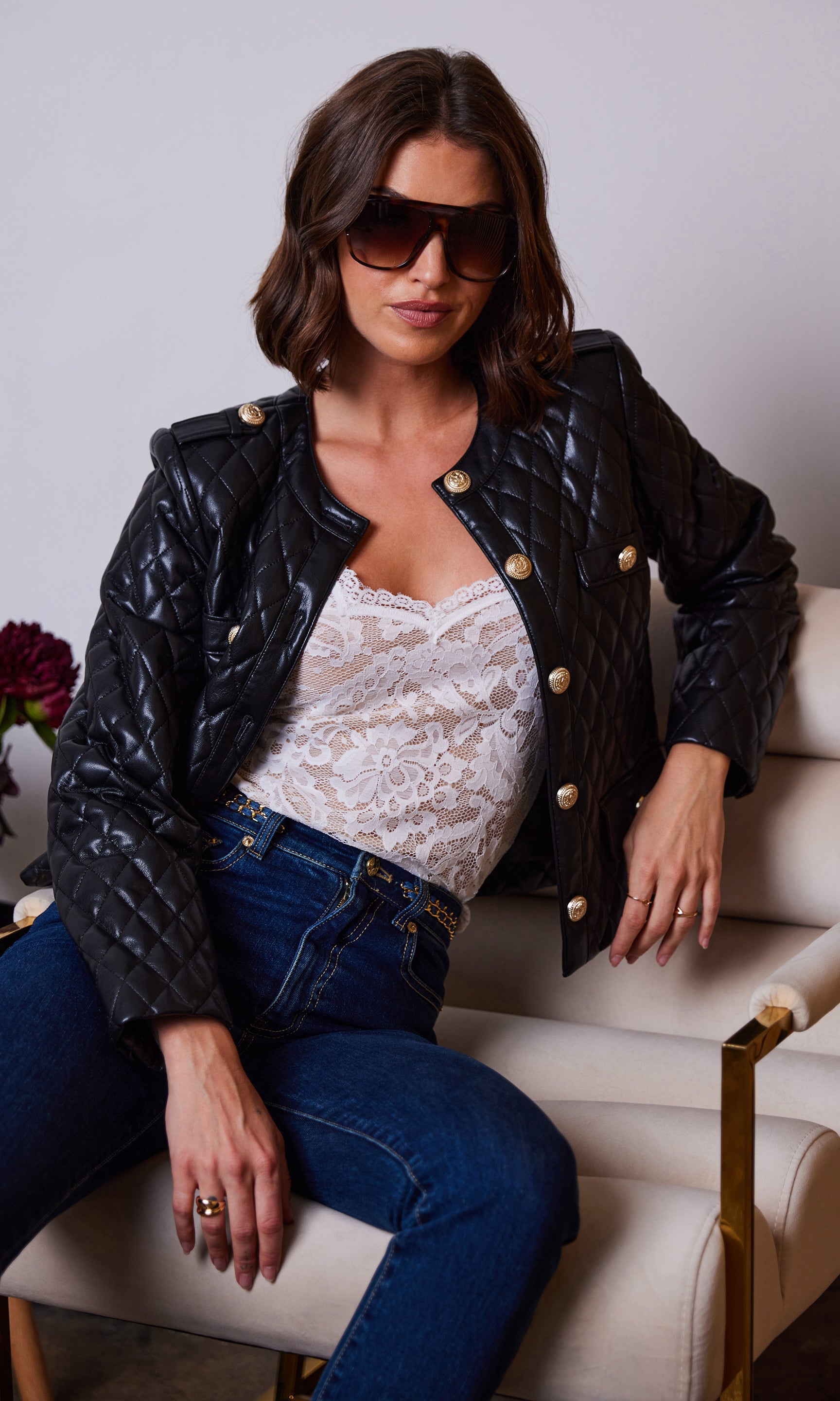 The model sits in a three-quarter shot wearing the Brevel Quilted Vegan Leather Jacket in black, leaning on a sofa to emphasize the jacket’s luxe sheen.
