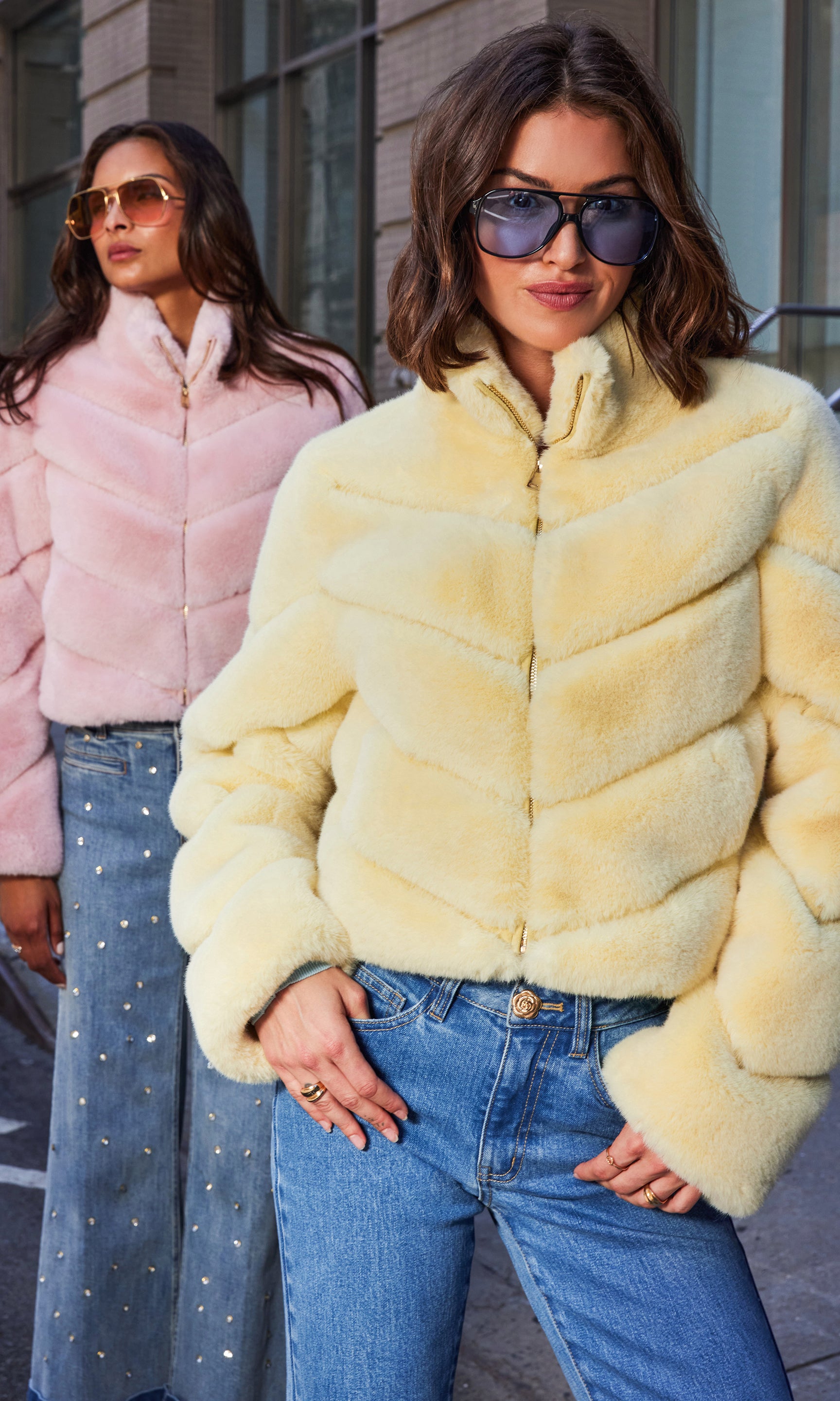 The models appear in a mid shot front view wearing the Brianna Faux Fur Jacket in banana color and ballet slipper, drawing attention to the plush chevron quilting and high collar under crisp urban light.
