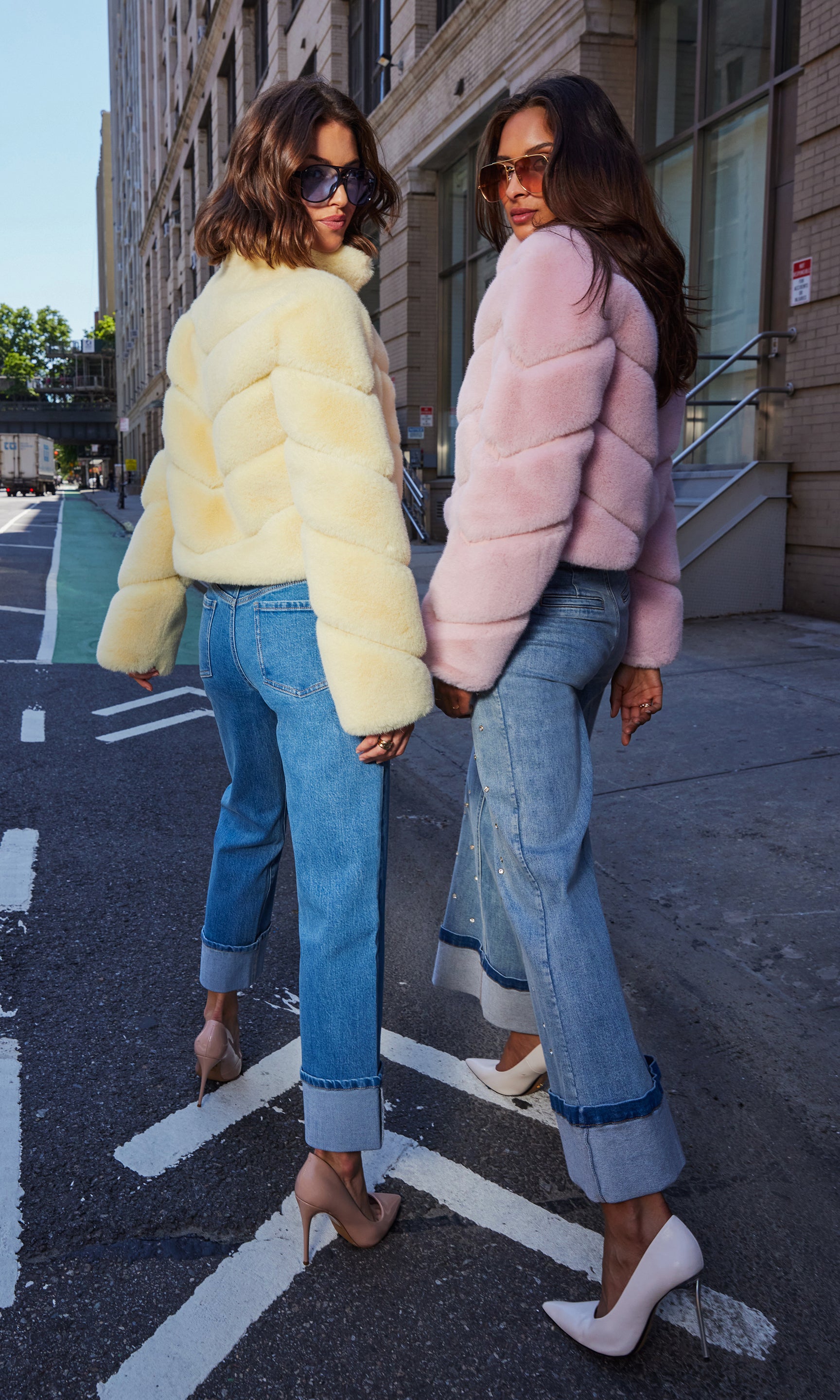 The models pose in a rear view wearing the Brianna Faux Fur Jacket in banana color and ballet slipper, highlighting their plush chevron quilting and cropped silhouettes against an urban street backdrop.