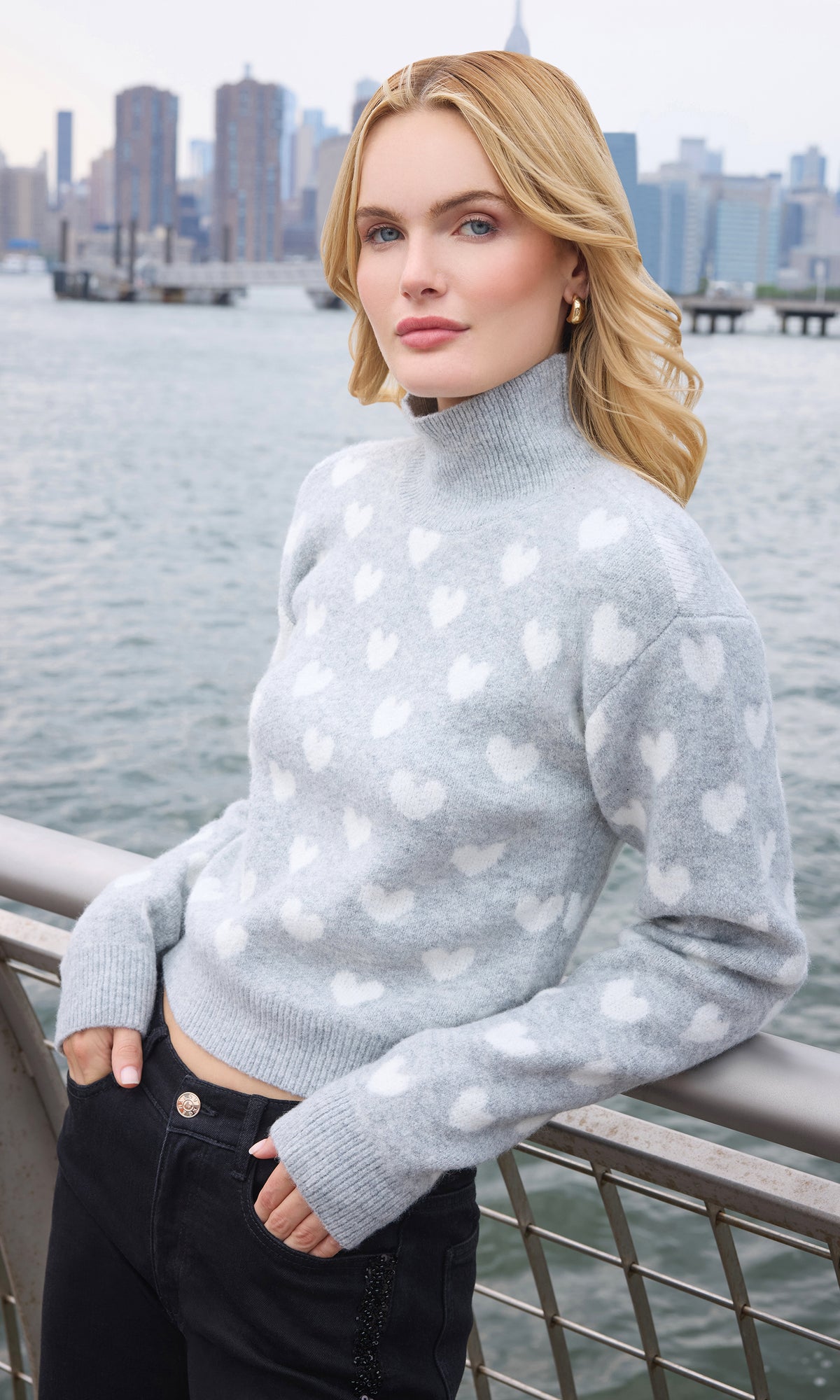 A blonde woman wearing a light gray mock-neck knit sweater with a white heart pattern, paired with black pants, standing on a balcony with a city skyline and water in the background.
