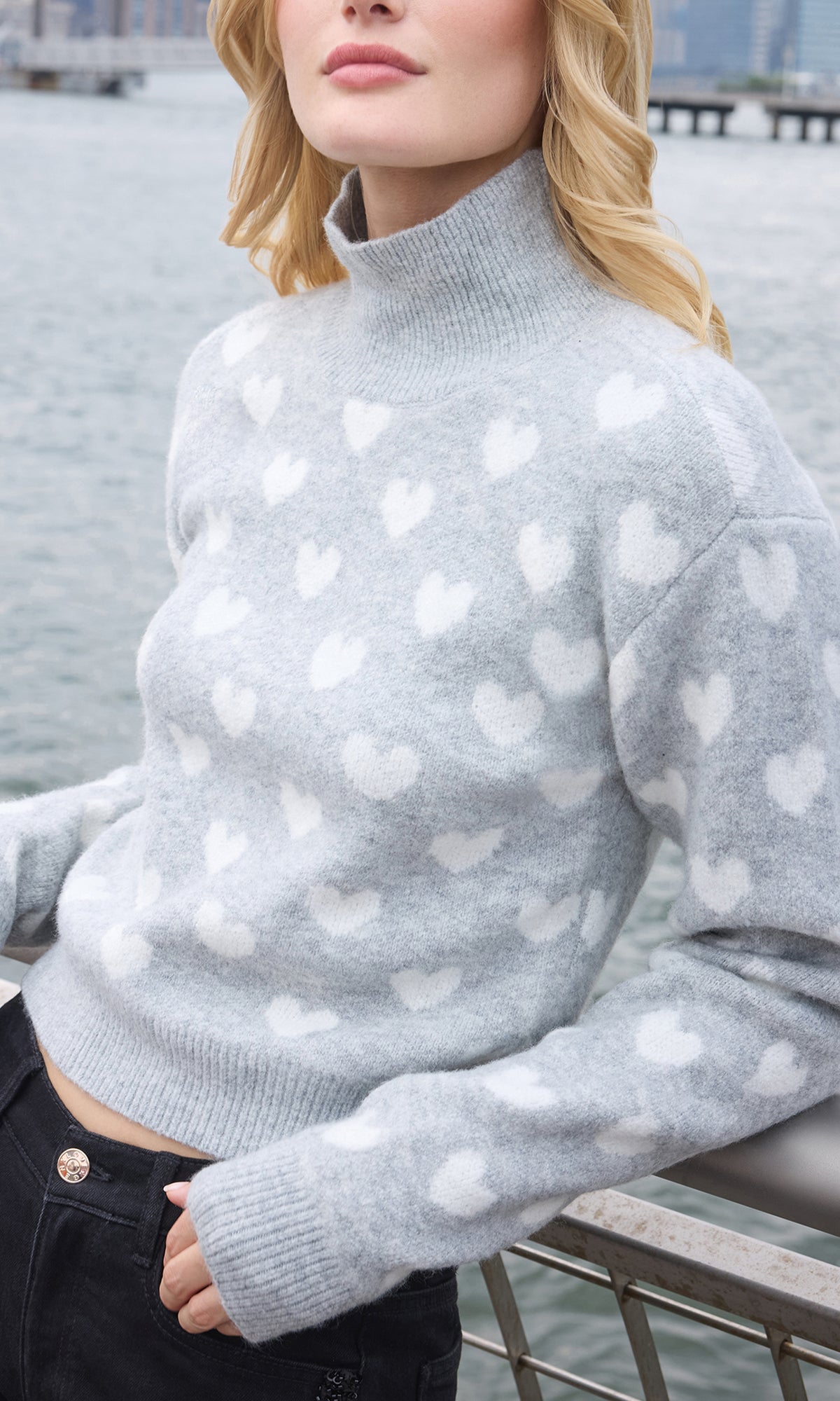 A woman wearing a light gray mock neck knit sweater with a white heart pattern, photographed outdoors near the water with a railing in the background.
