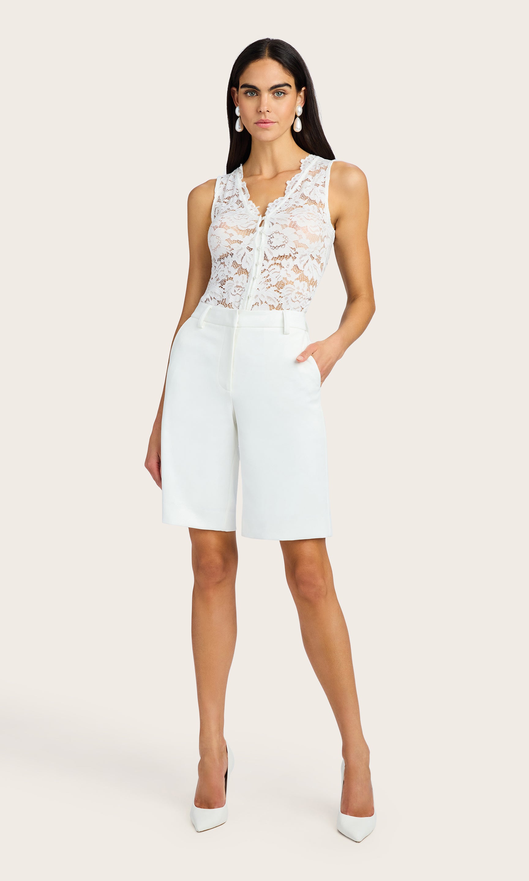 full body image of model wearing the Brinley lace tank in white with petite pearl buttons down the front, paired with white bermuda shorts and accessorized with big pearl rain drop earrings and white kitten heels
