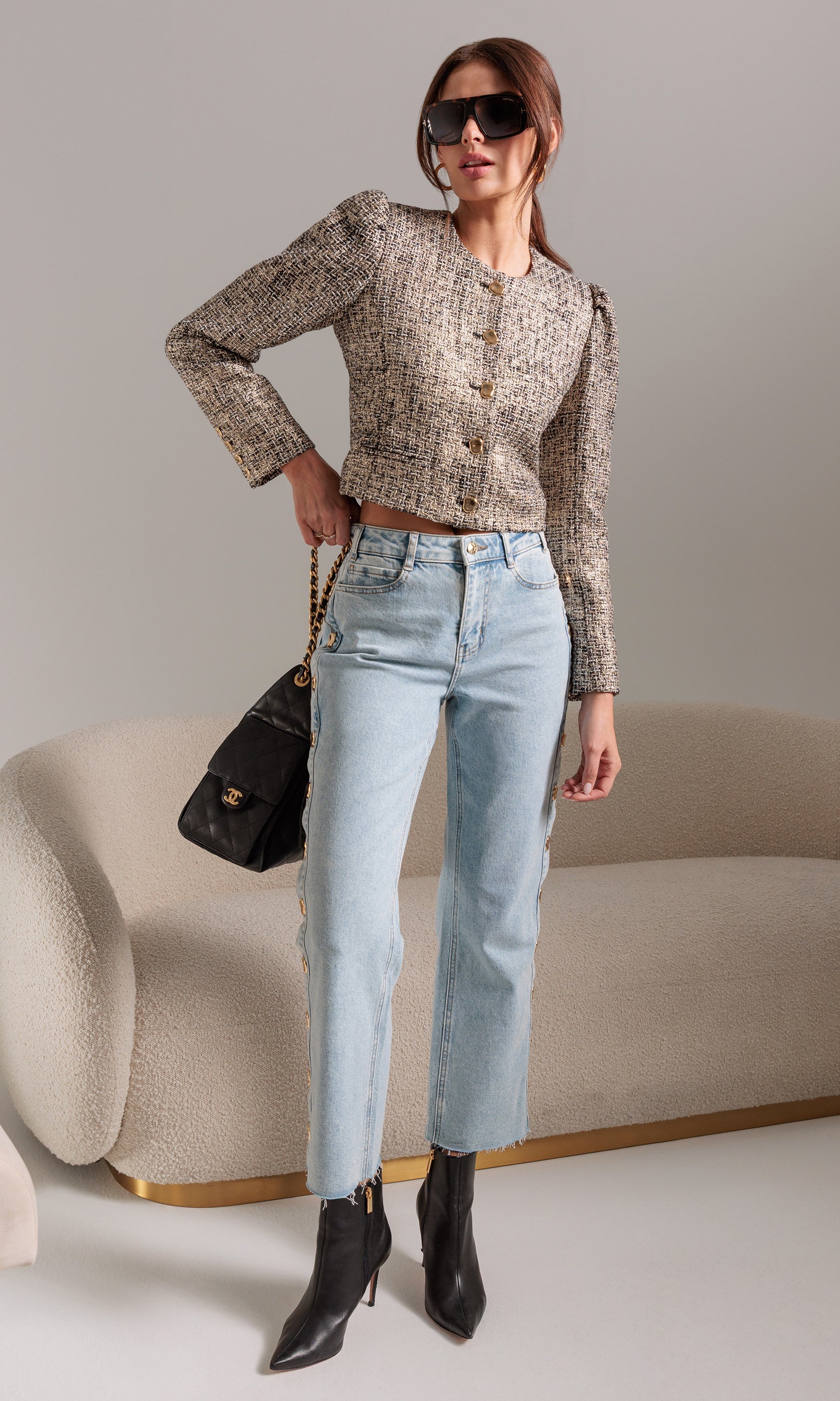 Woman wearing a patterned tweed jacket, light blue jeans, and black boots against a neutral background.