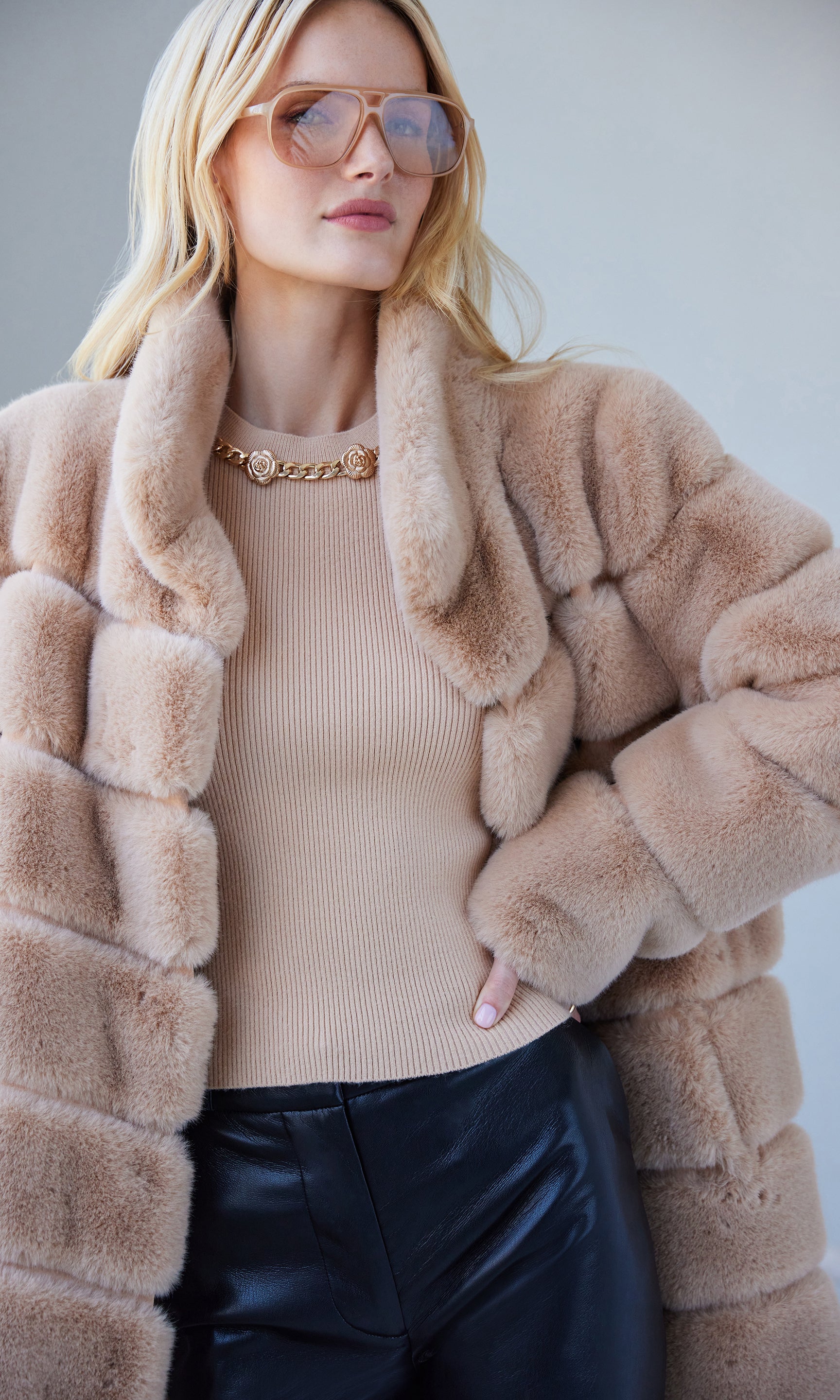 Woman wearing a beige fur coat, beige sweater, and black pants against a light background