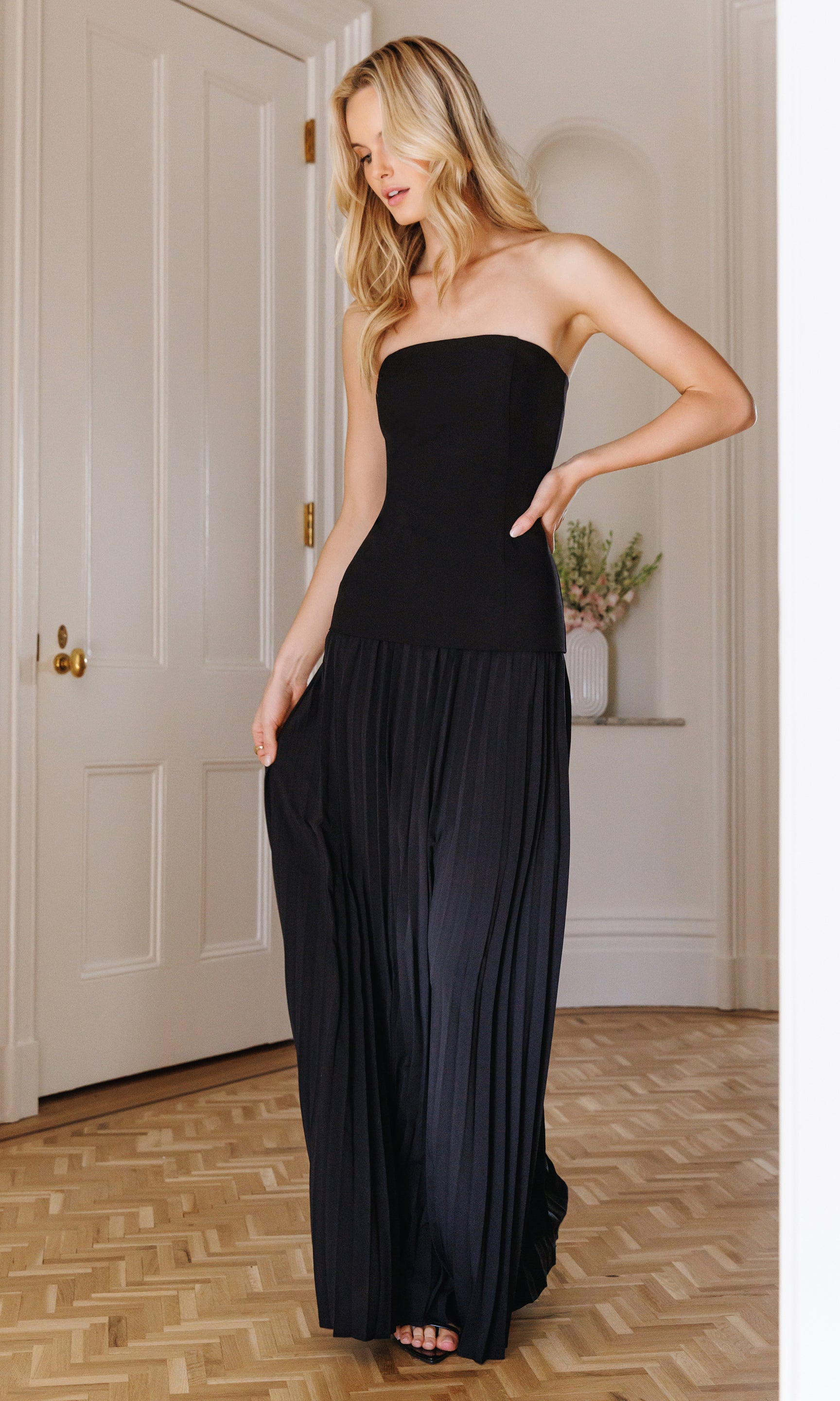 Model wears the Cody Crepe Pleat Dress in black, full body angled view showing the floor-length pleats and elegant drape from the side.