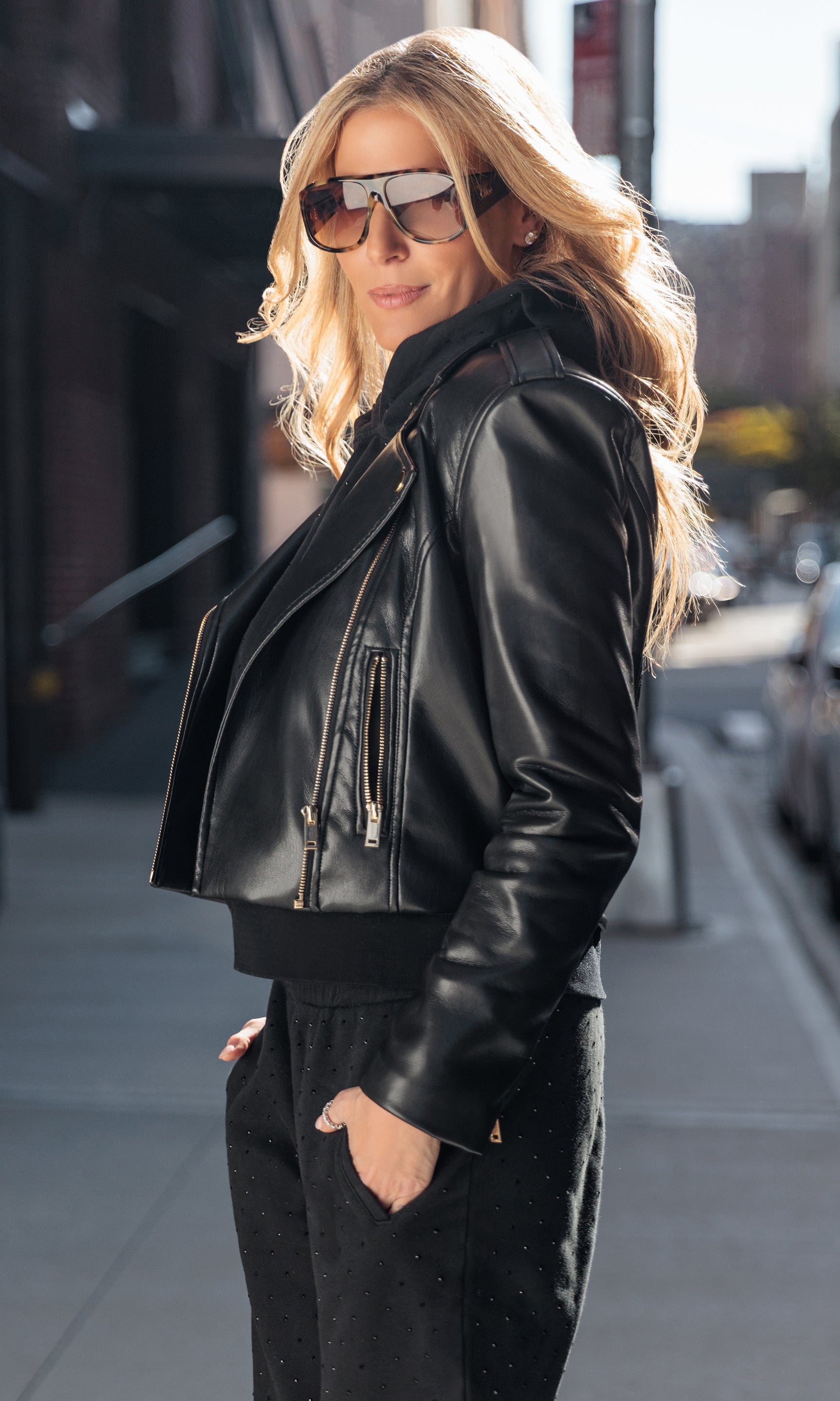 Woman wearing a black leather jacket and sunglasses on a city street.