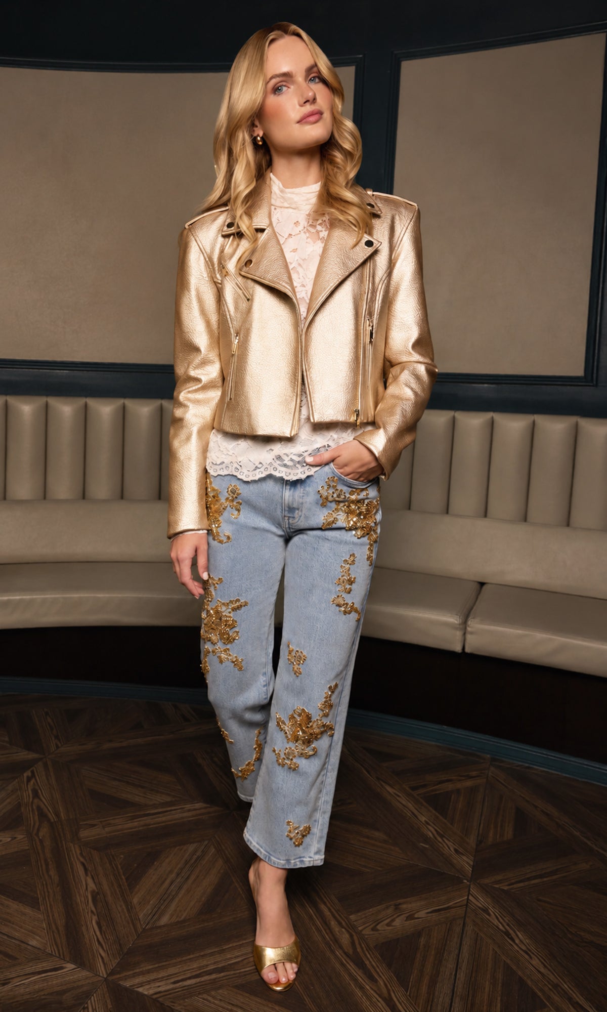 Model in an indoor setting wearing a gold moto jacket over a white lace top and blue jeans with gold floral applique, paired with gold heels.