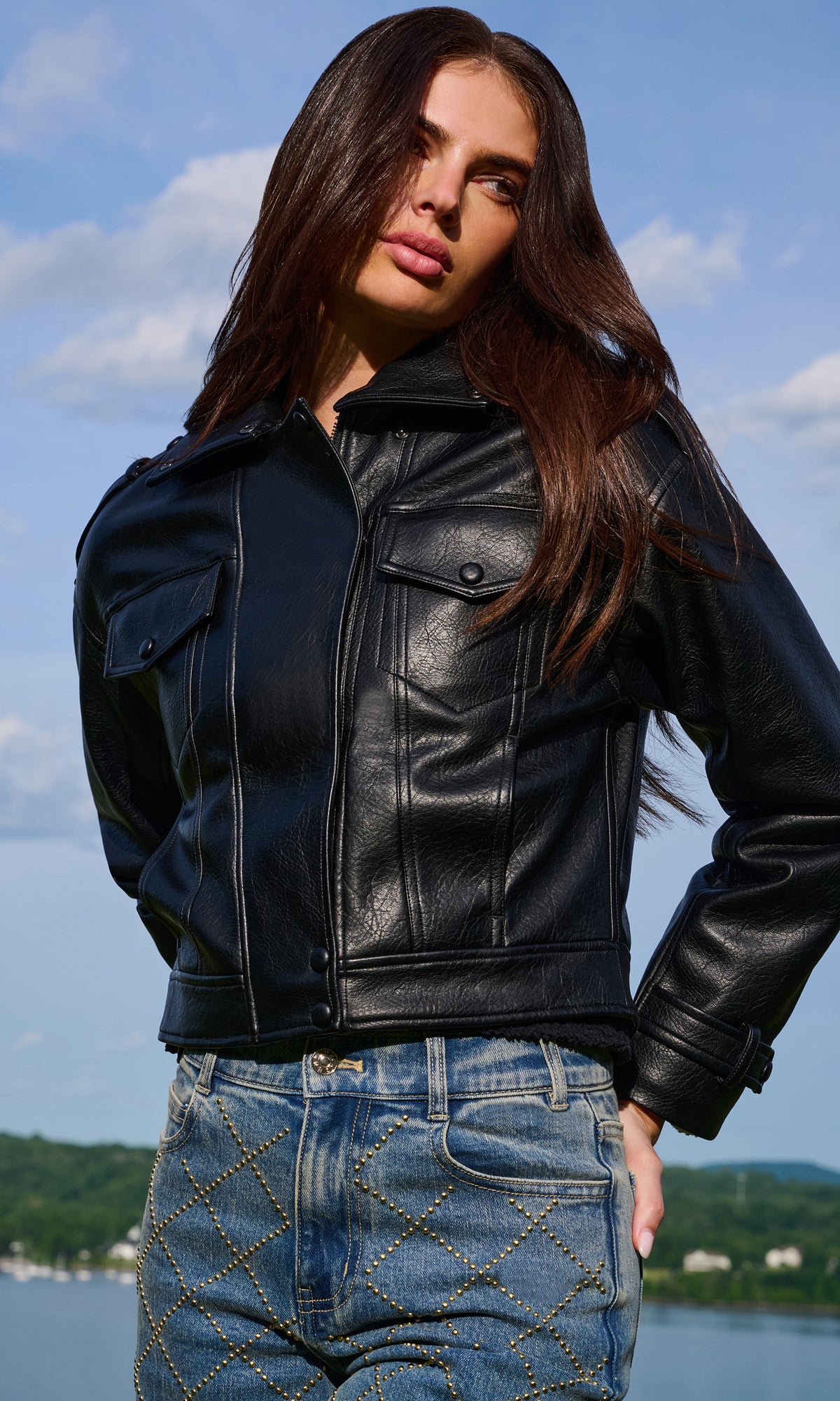 A woman wearing a black leather moto jacket and blue jeans with gold chain stitching, posing outdoors against a background of water and sky.
