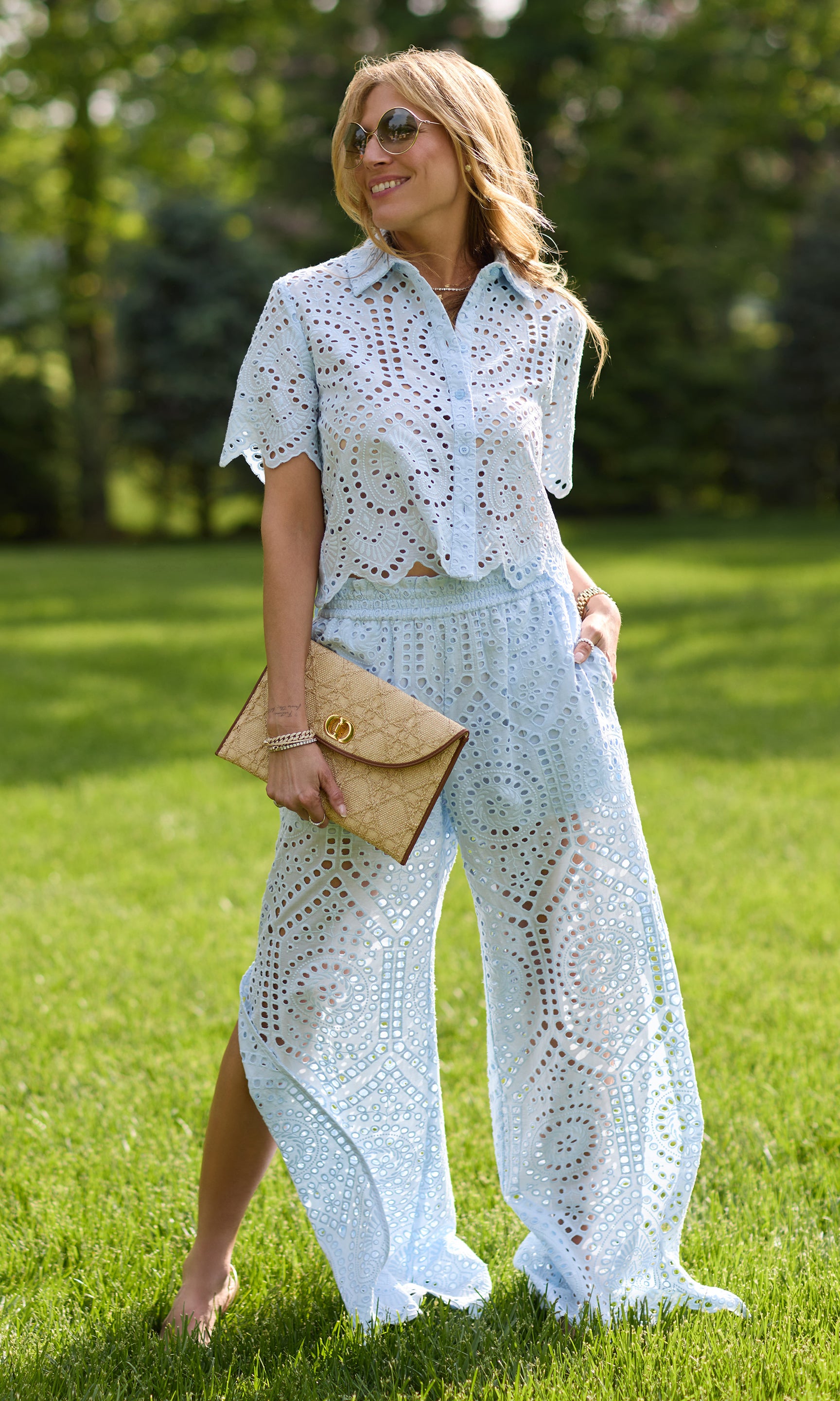 image of our founder Roni posing in a grass field wearing the blue cotton eyelet top and trousers set while holding a large brown clutch purse