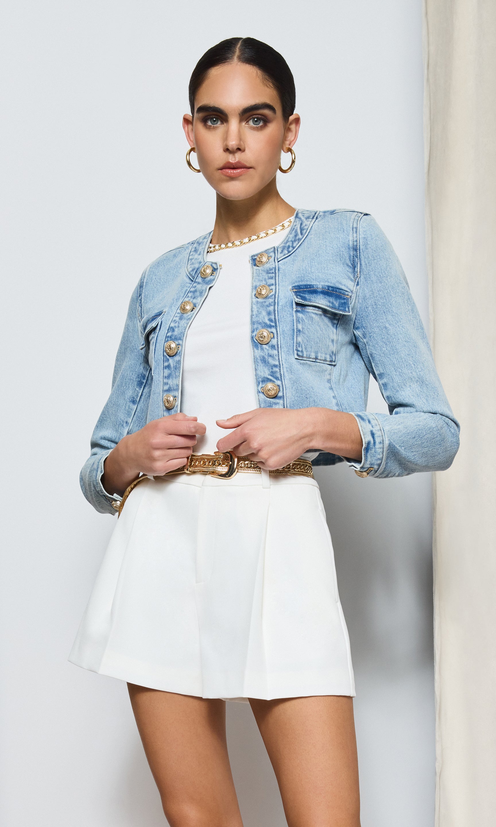 image of model standing in front of a beige curtain, wearing the Valentina denim jacket over a white romper