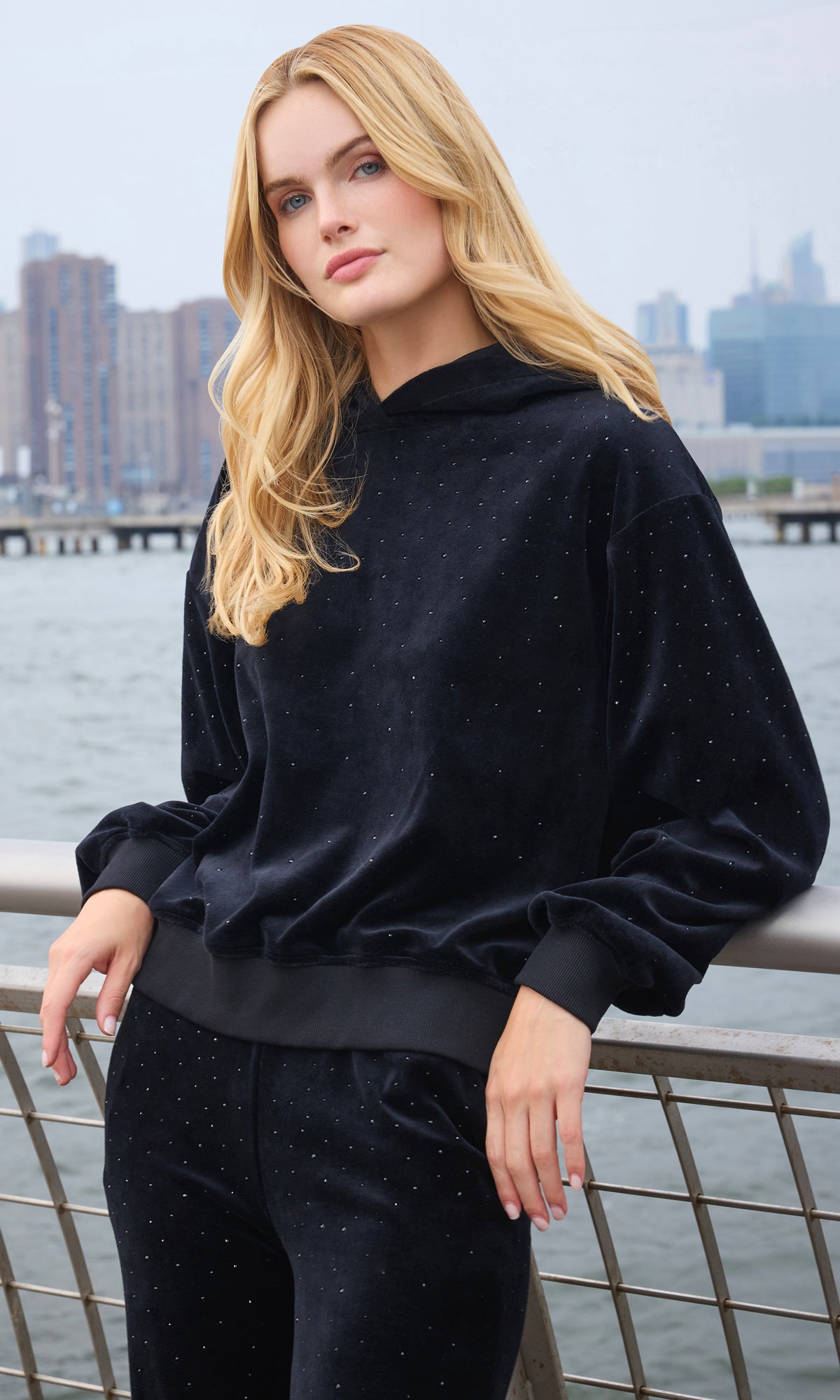 A blonde woman wearing a black velour hoodie and matching pants with subtle silver glitter speckles, posing outdoors with a city skyline and water in the background.