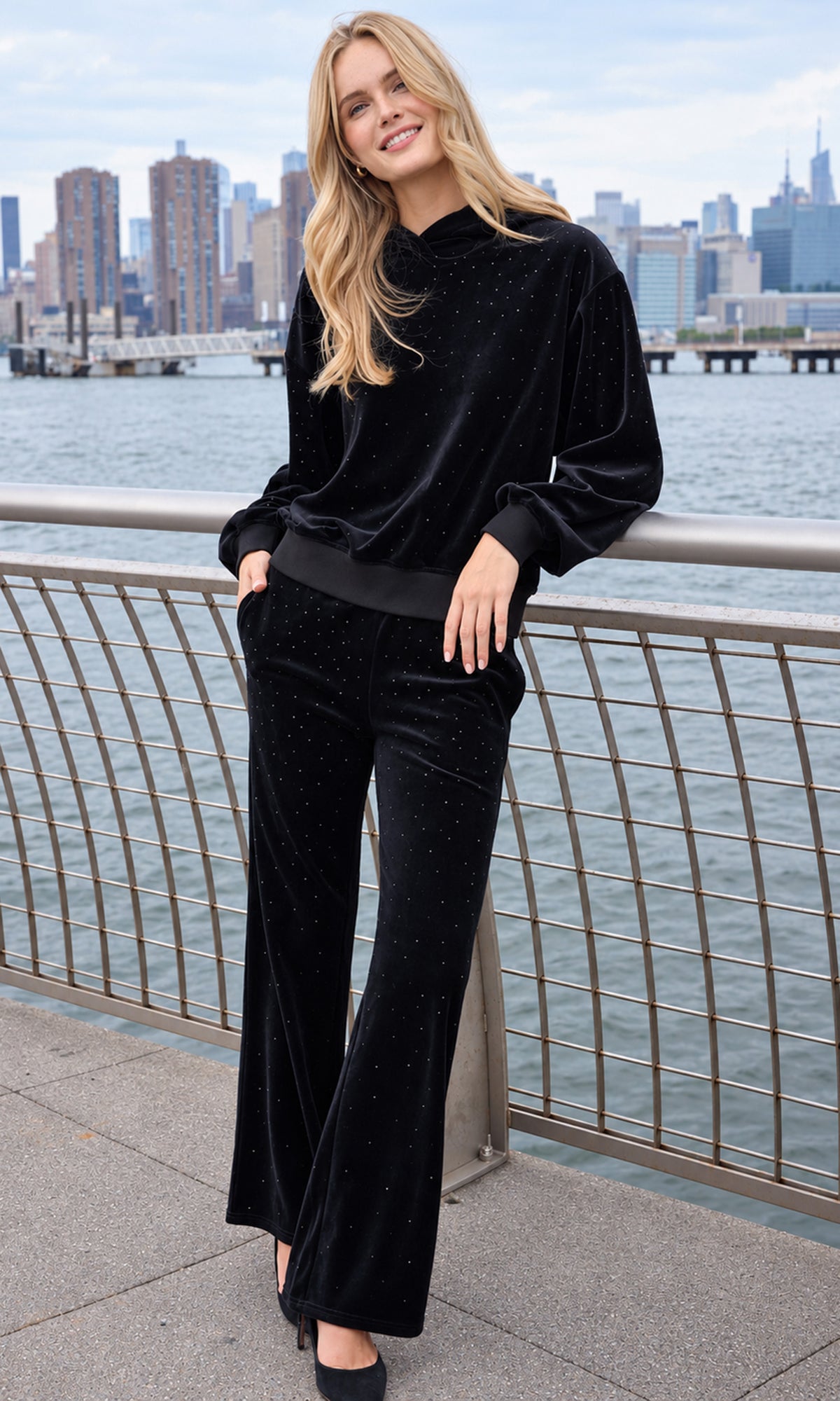 A woman stands outdoors by a waterfront railing wearing a matching black velvet two-piece outfit with a high-neck long-sleeve top and wide-leg pants.