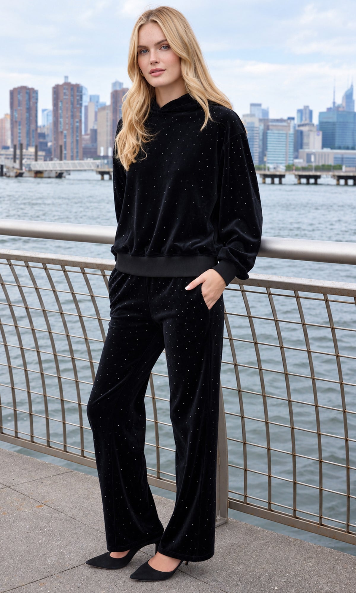 A woman stands outdoors by a waterfront railing wearing a matching black velvet two-piece outfit with subtle sparkly dots, black pointed flats, and a city skyline in the background.