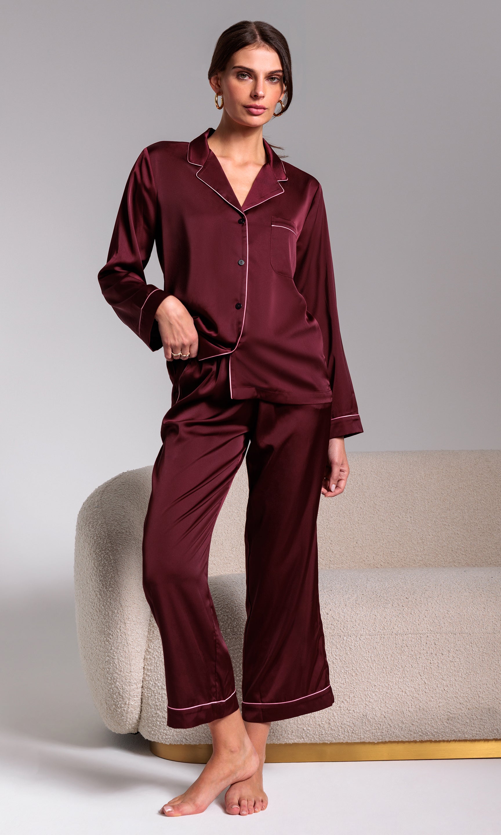 Woman wearing burgundy satin pajamas sitting on a beige chair against a gray background