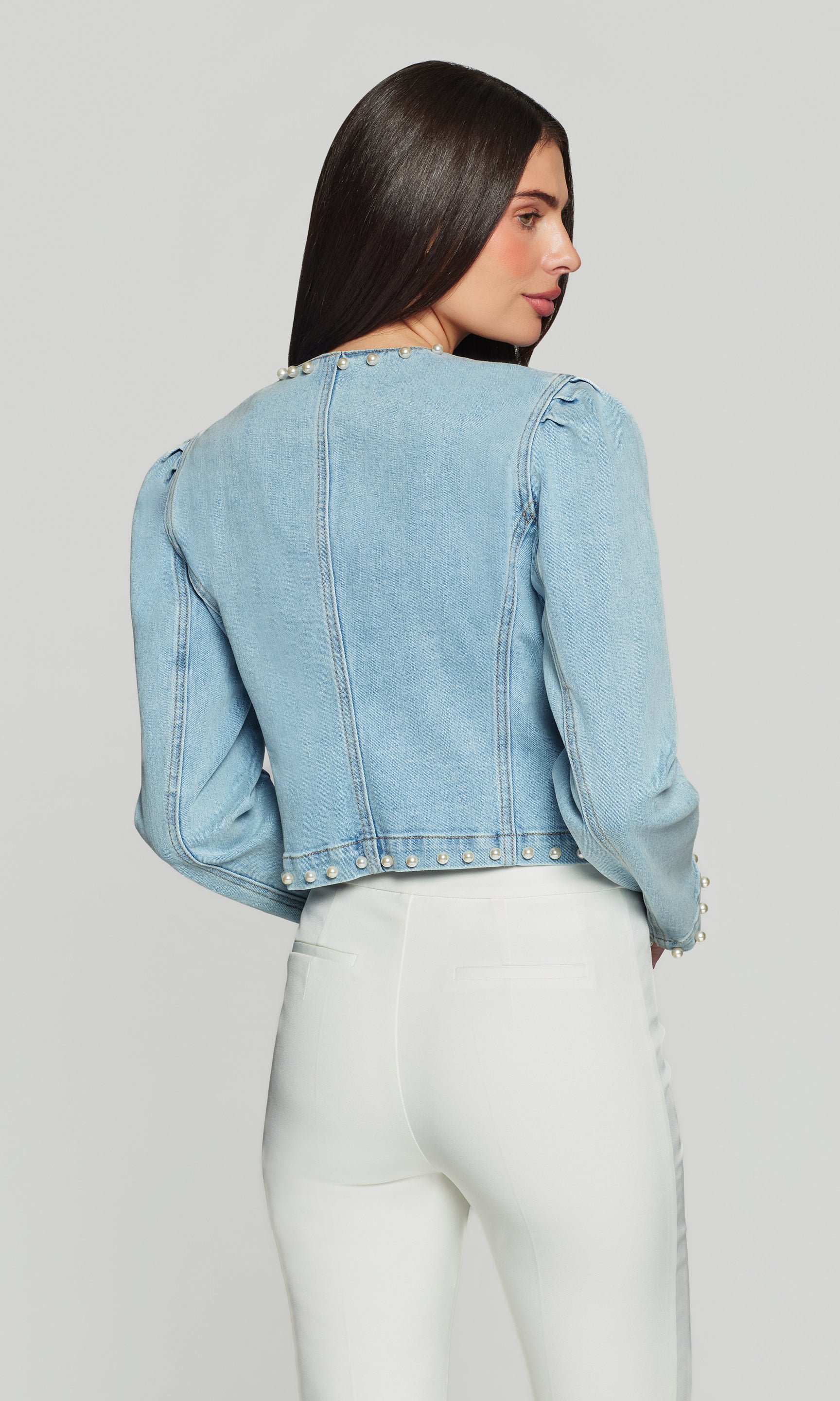 Back view of woman wearing a light blue denim jacket and white pants on a gray background