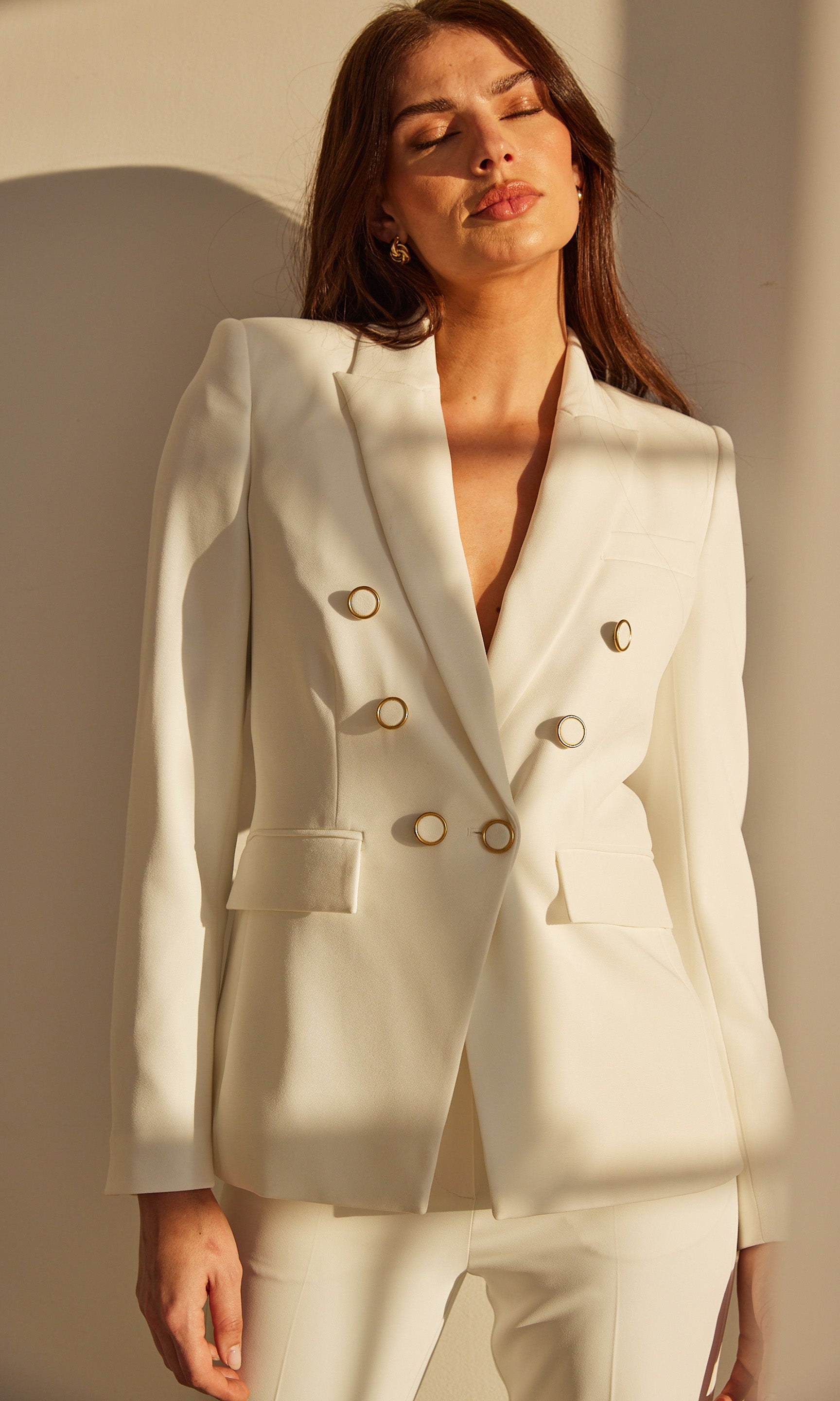 Indoor image of a model in a white double-breasted blazer and matching pants, softly lit setting.