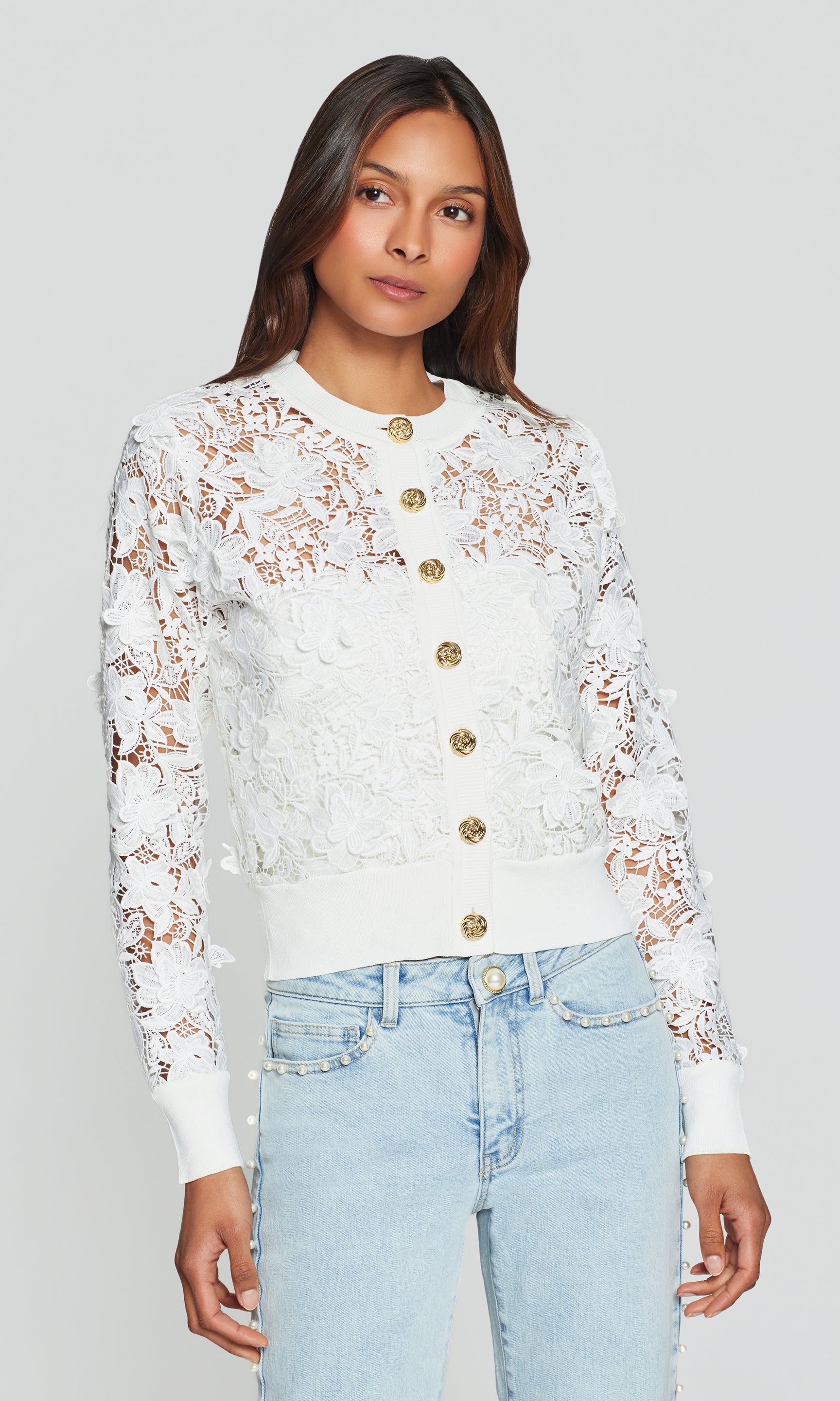 Woman wearing a white lace cardigan with gold buttons and light blue jeans on a gray background