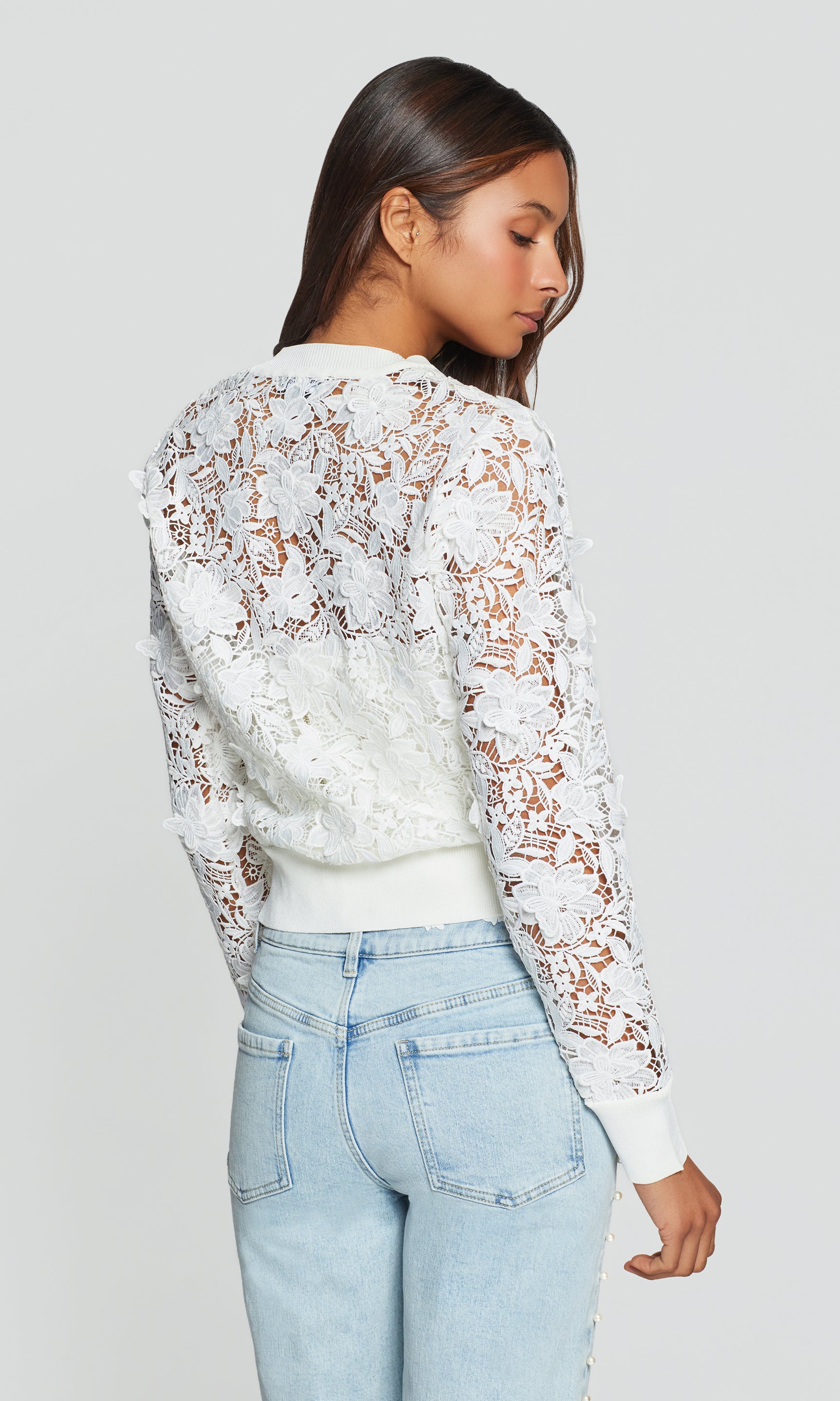Woman wearing a white lace long-sleeve cardigan and light blue jeans on a gray background
