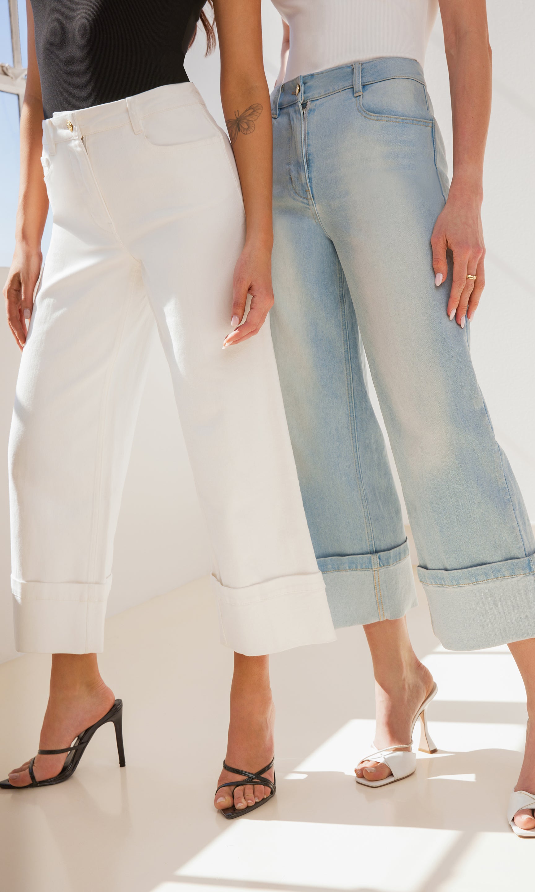 image of two models standing by each other in front of a window, both wearing the Eros jeans in different colors, both are paired with black shirts and black heels