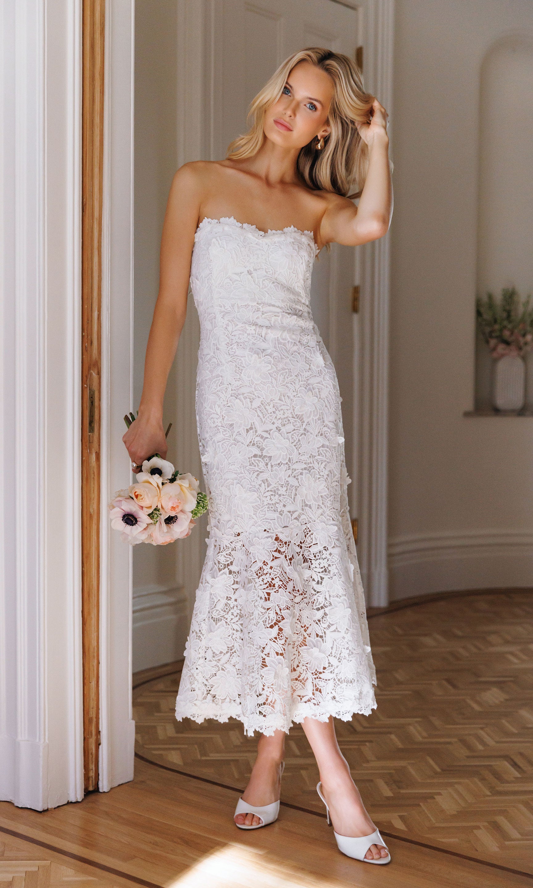 Woman in an indoor setting wearing a white lace dress holding a white floral bouquet.